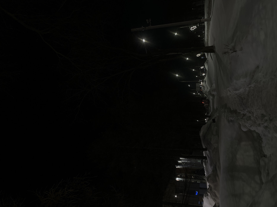 This nighttime image captures a snow-laden urban environment in Chaikovsky, Russia, during winter. A cleared, trodden pedestrian path, marked by footprints, traverses diagonally from the lower foreground into the midground, flanked by significant accumulations of undisturbed snow. The scene is primarily illuminated by numerous bright, white streetlights mounted on utility poles, casting light onto the snow and parts of the road. On the far left, a multi-story building with illuminated windows is partially visible. To the right of the pedestrian path, a road is discernable in the midground, with faint red vehicle lights in the distance. Several white, snowflake-shaped light decorations are affixed to poles along the right side of the road. Bare deciduous trees, devoid of leaves, are silhouetted against the dark sky and stand near the path and road. No individuals are present.
