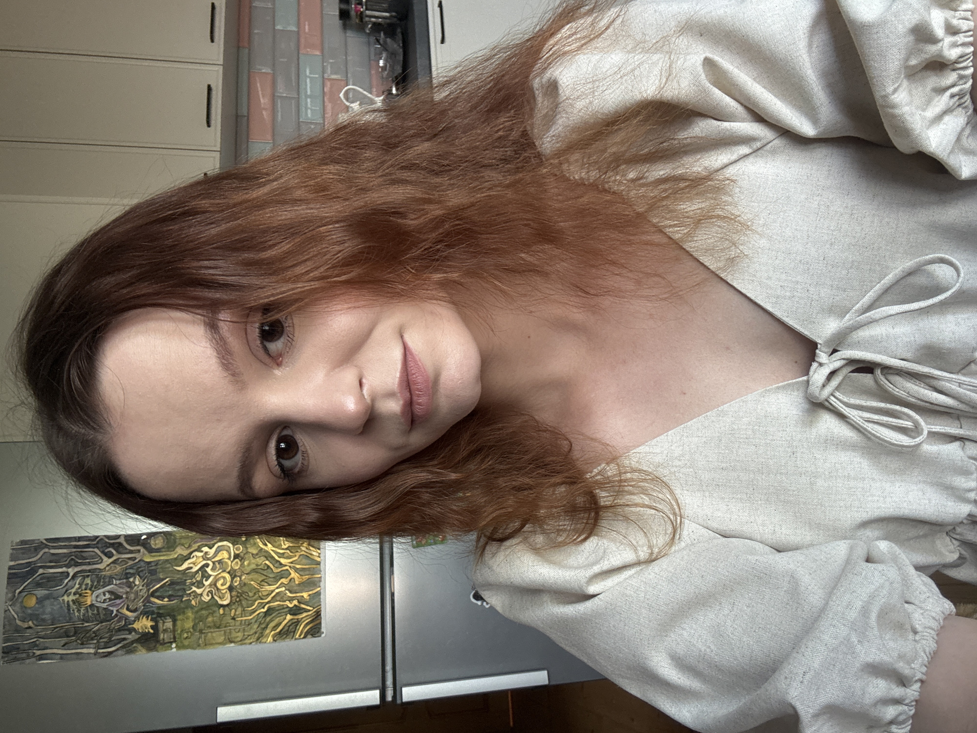 A young woman with reddish-brown hair, fair skin, and brown eyes is looking directly at the camera. She is wearing a light-colored, V-neck top with puffy sleeves that are cinched at the wrist. The top has a tie closure at the neckline.
The background includes a silver refrigerator with a framed piece of art hanging on it. The artwork depicts a stylized figure in a forest setting with flames. To the right of the refrigerator, kitchen cabinets and a tiled backsplash are visible. The overall setting appears to be an indoor domestic environment. There is no indication of the specific location within the city of Rzhev, Russia.