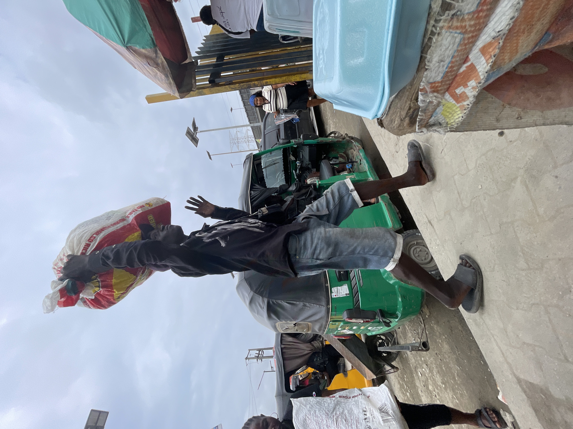 In Ajah, Nigeria, a young man carries a large bag of rice over his head. He is dressed in a black hoodie and denim shorts, with black flip-flops on his feet. The bag is red, yellow, and white, and appears to be a branded product. He is walking on a paved area, with a green auto-rickshaw behind him. In the background, there are more auto-rickshaws, a woman carrying a white bag, and a stall with a blue plastic container. The sky is overcast, suggesting it might rain. The scene appears to be a busy market area.