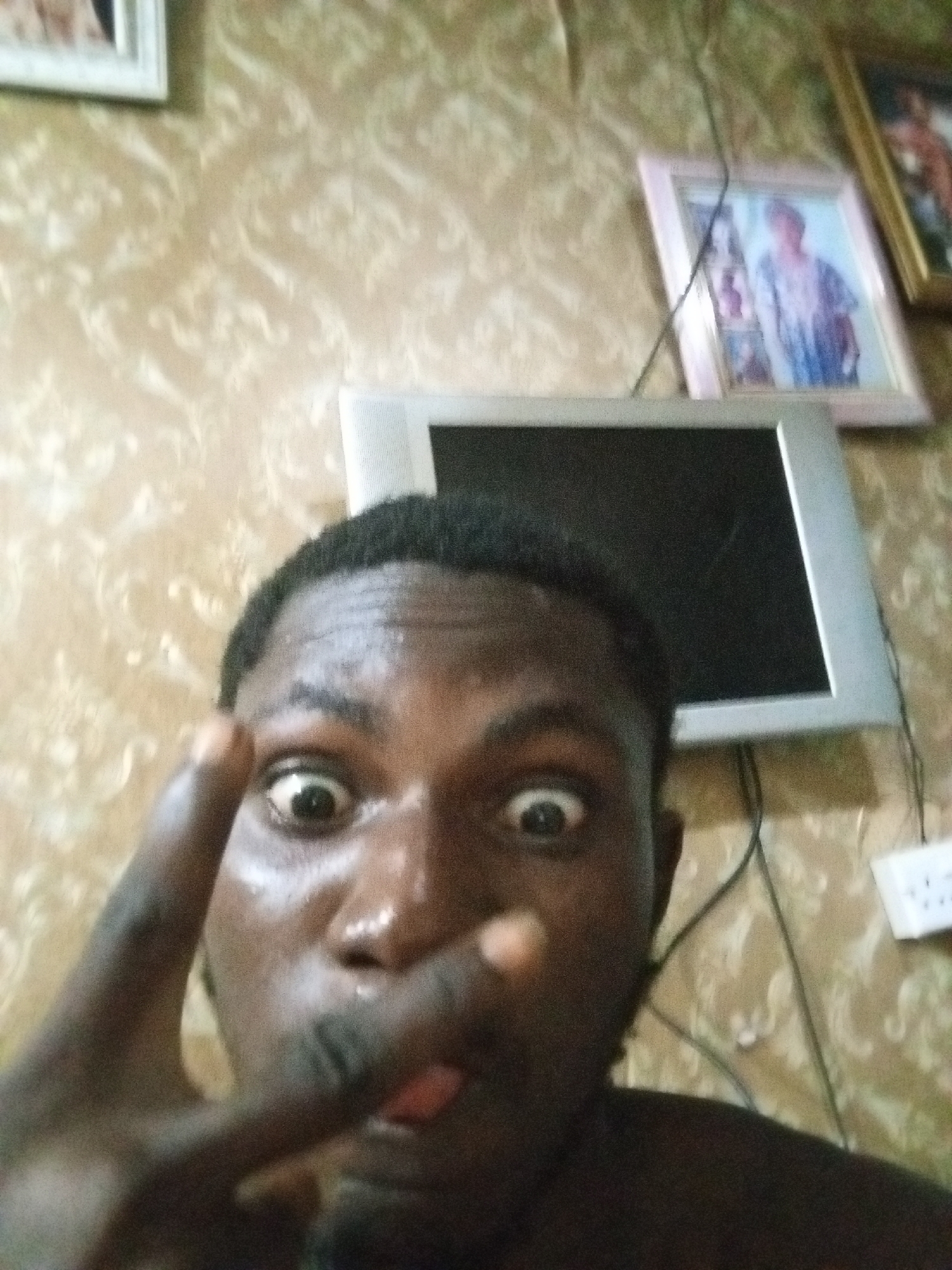 The image depicts a person's face in close-up, making a face with wide eyes and sticking out their tongue. The person is also holding up their fingers in a peace sign. Behind the person, the wall has a beige patterned wallpaper. There is a silver-framed monitor and two framed pictures hanging on the wall. A white electrical outlet is also visible. The location is identified as Unknown, Unknown.