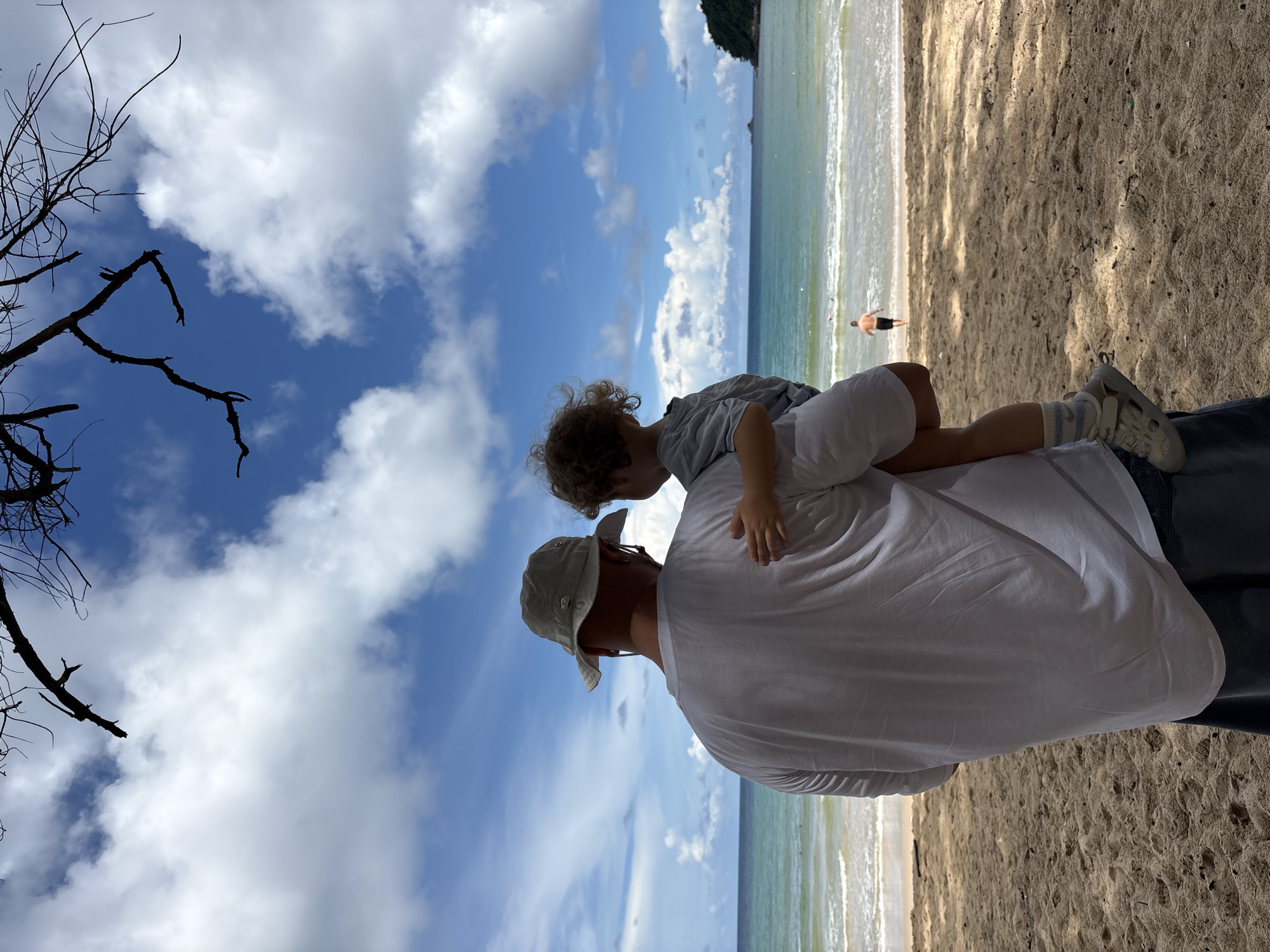 A man wearing a bucket hat and a white t-shirt is carrying a young child on a sandy beach. The child has curly brown hair and is wearing a grey t-shirt. They are facing away from the camera, looking out at the ocean. The sea is a clear blue-green, with small waves breaking on the shore. In the distance, a small island with lush green vegetation is visible. A tree branch with a few leaves hangs from the top of the frame. The sky is bright blue with scattered white clouds. The sand on the beach is light brown and appears dry. Another person, a man wearing dark shorts, is seen walking into the water in the distance. The overall scene is one of a peaceful day at the beach in Choeng Thale, Thailand.