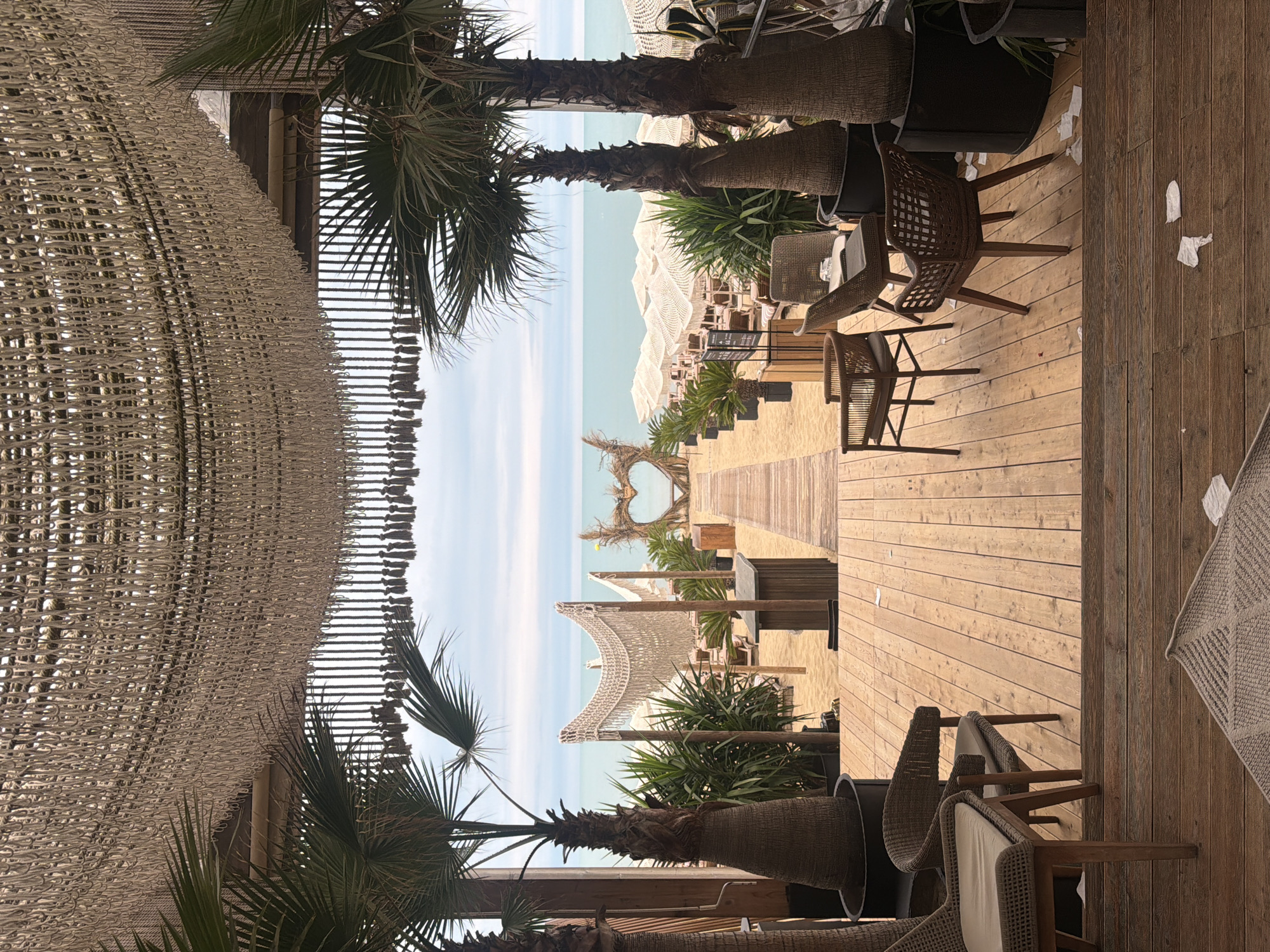 This image depicts a sun-drenched, outdoor beachside seating area, likely a restaurant or cafe, with a clear view of the ocean. The scene is dominated by a wooden deck furnished with several chairs, some made of woven material and others with upholstered seats. Palm trees are scattered throughout the area, adding to the tropical ambiance.
In the foreground, to the left, a large structure covered in a woven netting, possibly a shade sail or decorative element, hangs overhead, partially obscuring a palm tree. Beyond this, the turquoise ocean stretches towards the horizon, meeting a pale blue sky. The beach itself is visible, lined with rows of white lounge chairs and umbrellas, suggesting a resort or beach club setting.
A prominent feature in the middle ground is a heart-shaped archway made of branches and foliage, positioned on the sand and leading towards the sea. Wooden pathways guide visitors across the sand. The overall atmosphere is relaxed and inviting, indicative of a pleasant day at the beach. The lighting suggests it is daytime, with bright sunlight casting shadows. No people are actively visible in the frame, contributing to a sense of tranquility. The image conveys a feeling of a serene vacation or leisurely day outdoors.
