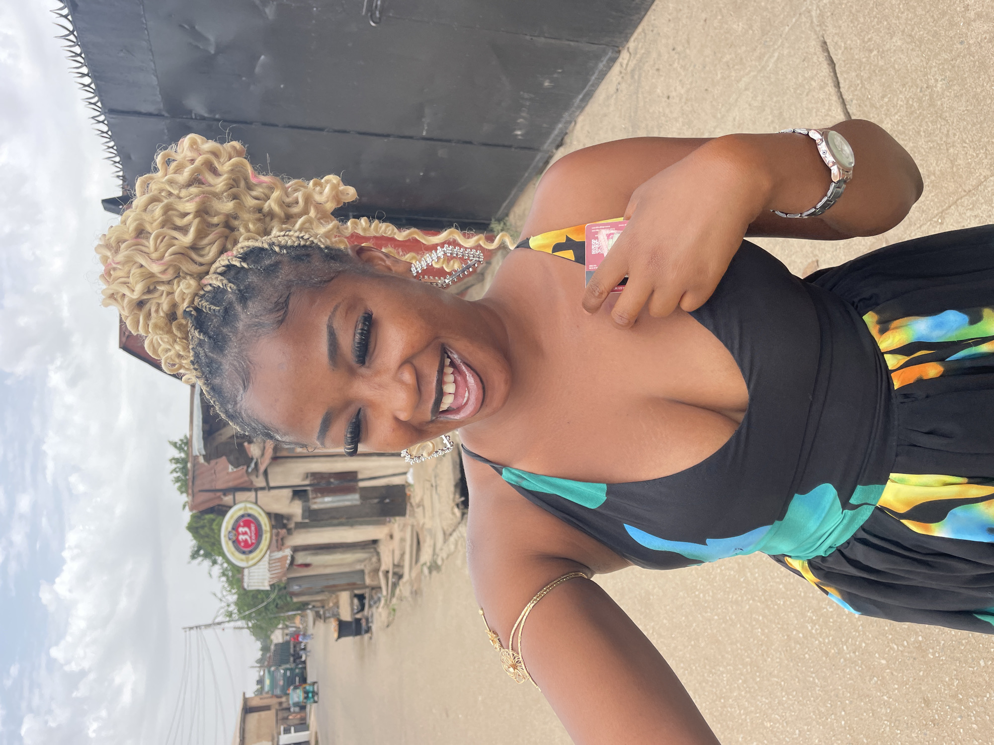 A woman smiles and holds a small card with colorful designs in her left hand. She is wearing a black dress with colorful teal, blue, yellow, and orange accents. The woman has blonde, curly, braided hair, and is wearing silver earrings and a silver watch on her right wrist. She also has a gold arm cuff on her left arm. The photo appears to be taken in Jos, Nigeria, as evidenced by a sign for a “33” Export beer, a popular Nigerian brand, visible in the background street. The background shows a street with buildings and cars.
