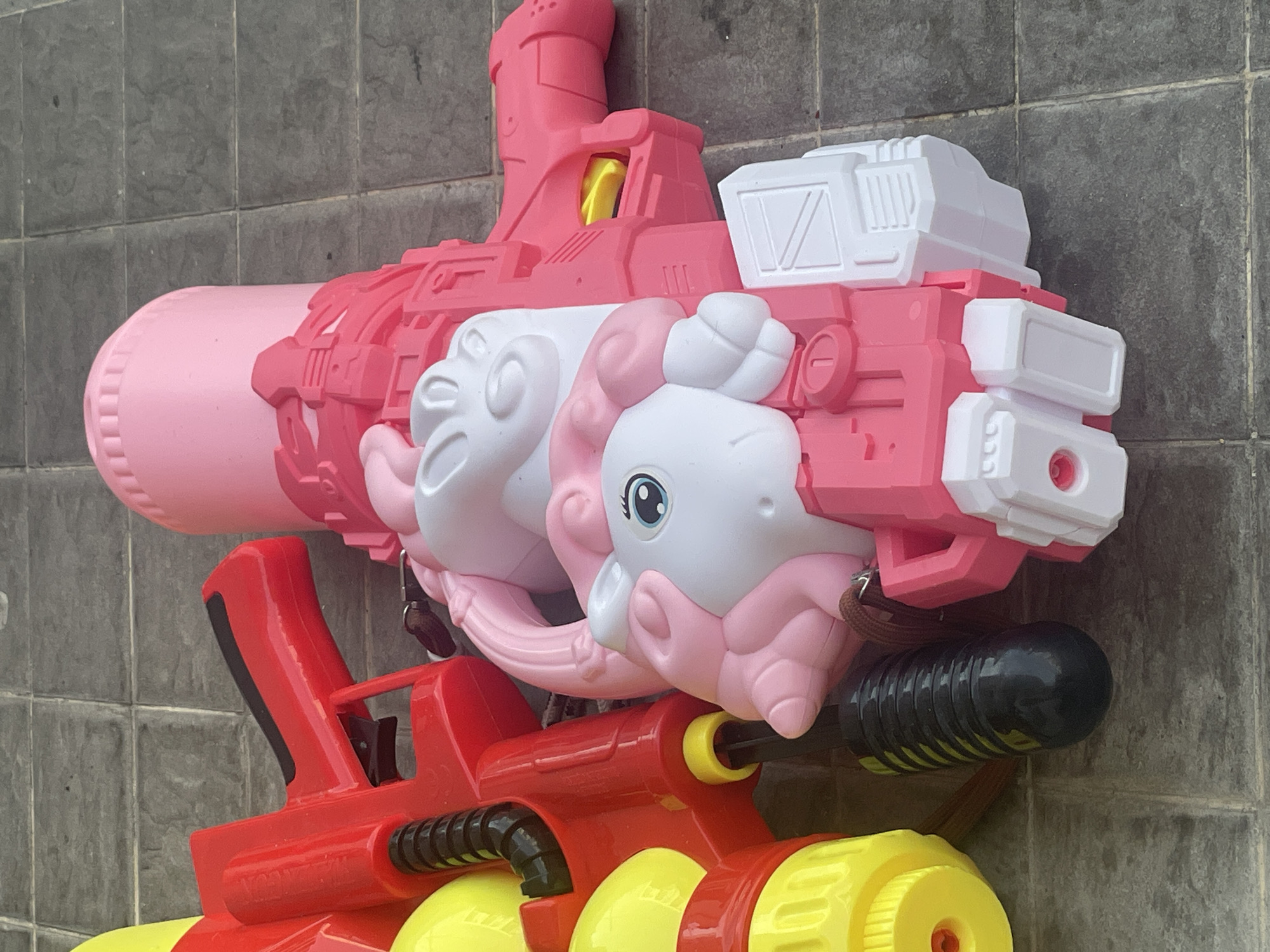 Two toy water guns are positioned against a gray tiled wall. The top water gun is pink and white, featuring a unicorn design. The lower water gun is red and yellow. The location is Chang Phueak, Thailand.