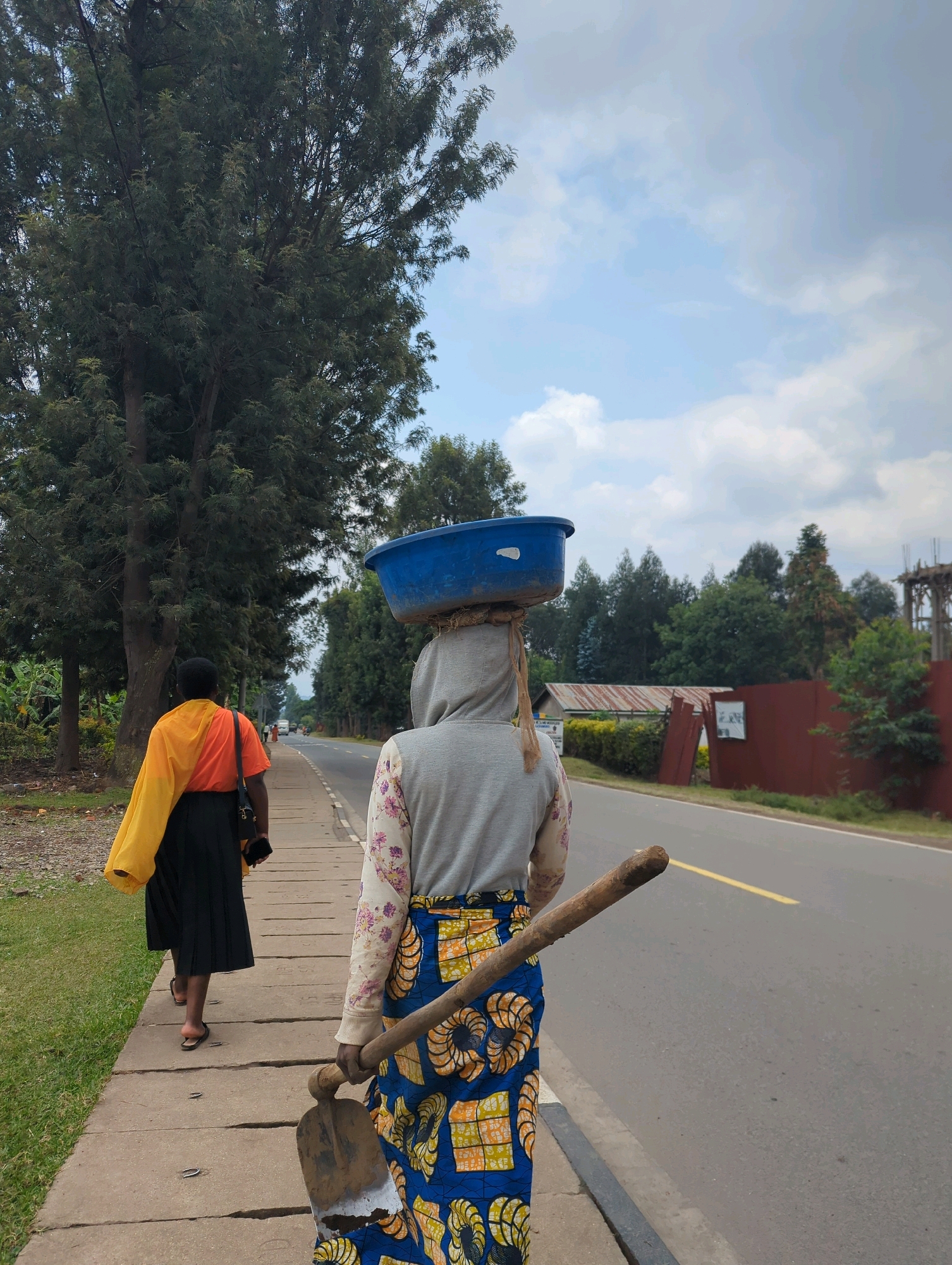 Two individuals are walking on a paved sidewalk next to a two-lane asphalt road. The person in the foreground, facing away from the camera, is wearing a grey hooded sweatshirt with a floral-patterned sleeve and a colorful patterned skirt. They are carrying a hoe with a wooden handle over their right shoulder and have a blue plastic basin balanced on their head. The second person, also facing away, is further down the sidewalk and wears an orange shirt with a yellow cloth draped over their left shoulder and a black skirt. They appear to be holding a black smartphone in their left hand. The environment is characterized by lush green trees and vegetation along the left side of the sidewalk, with a rural landscape visible in the background, including a building with a corrugated metal roof and a partially constructed building with visible rebar. The sky is overcast with some patches of blue. The scene is set in Musanze District, Rwanda.