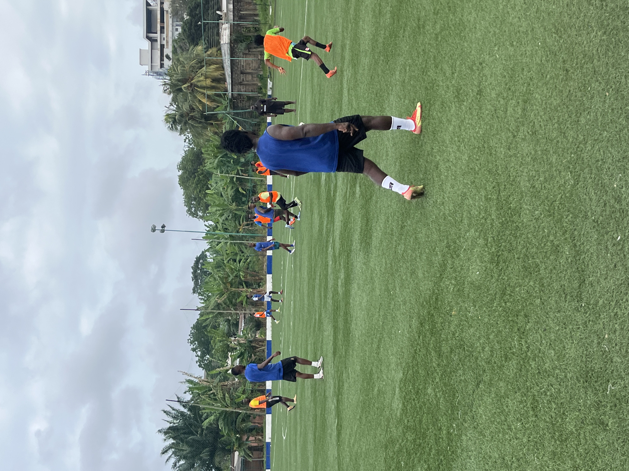 A group of male soccer players are practicing on an artificial turf field under an overcast sky. The field is marked with white lines and has a low blue and white striped fence surrounding it. In the background, there are palm trees, banana trees, and some buildings, including what appears to be a modern structure under construction on a hill.
The players are wearing various athletic attire, including blue, orange, and black shirts, and shorts. One player in the foreground is wearing a blue sleeveless jersey and black shorts, with white socks and brightly colored soccer cleats. Another player, further back on the right side of the frame, is wearing an orange vest over a black shirt and black shorts. Several other players are visible scattered across the field, engaged in drills or movement.
The overall impression is of an outdoor sports training session. The presence of lush tropical vegetation and the style of the buildings suggest a location in a warm climate, consistent with Accra, Ghana.
