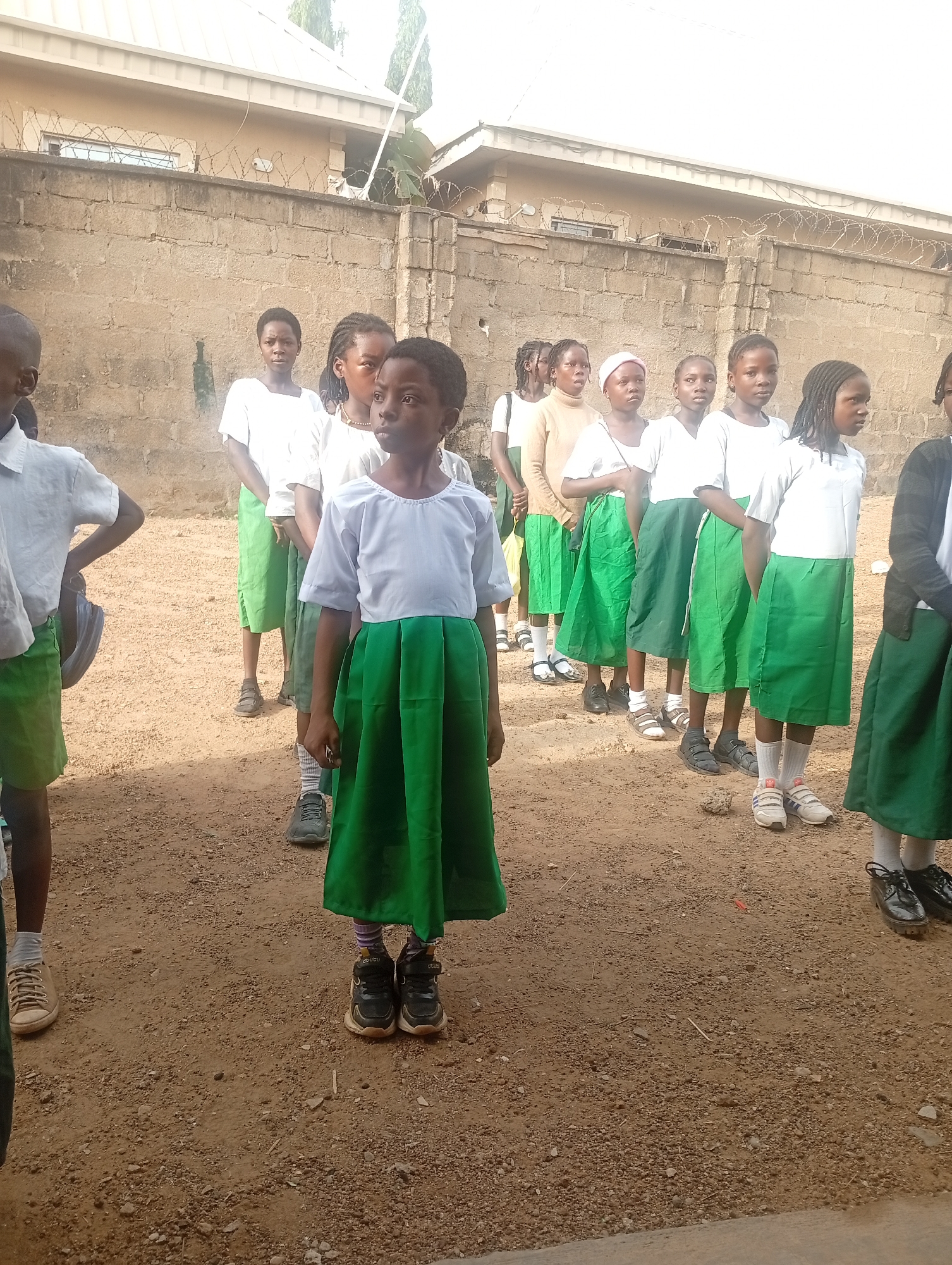 A group of young girls stand in a line on a dirt ground. They all wear a similar school uniform consisting of a white shirt and a green skirt. They are standing in front of a light brown wall that is topped with barbed wire. The location is Narayi, Nigeria.