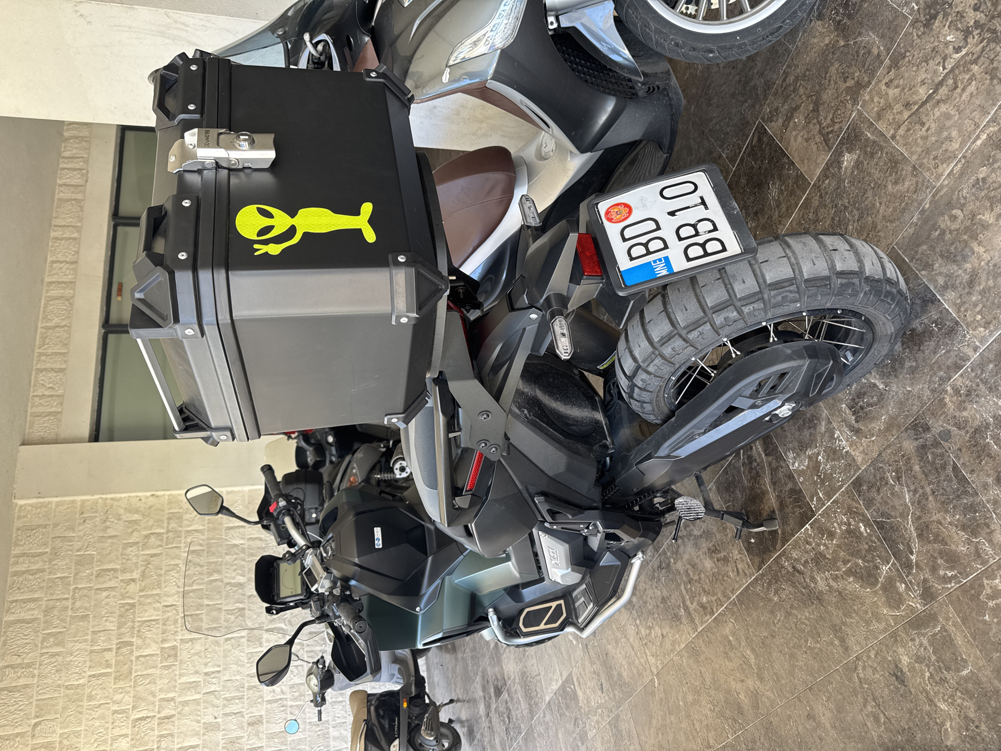This detailed image captures a rear-three-quarter view of a dark grey/green Honda X-ADV adventure scooter, parked on a tiled surface in Budva, Montenegro. The scooter features a large black top case secured with a silver metallic latch and black handles. A prominent, reflective neon yellow alien sticker, depicted with its left hand raised in a peace sign, adorns the left side of the top case. The Montenegrin license plate, clearly visible on the rear of the scooter, displays "MNE" for Montenegro, "BD" for the city of Budva, and the alphanumeric sequence "BB10." The scooter's rear wheel is fitted with robust, knobby tires suitable for off-road travel, and a black side stand is deployed.
To the left of the main scooter, a smaller, light brown-seated scooter is partially visible. Another silver-grey scooter, also equipped with a brown seat, is partially obscured to the right of the main subject. The vehicles rest on a floor composed of large, dark brown and grey mottled tiles with intricate lighter veins, resembling a natural stone pattern. The background features a textured, light-colored wall constructed from horizontally coursed bricks or stones. A light-colored vertical pillar stands on the right side of the frame, and further back, darker rectangular shapes suggest windows or doorways. No individuals are present in the scene.