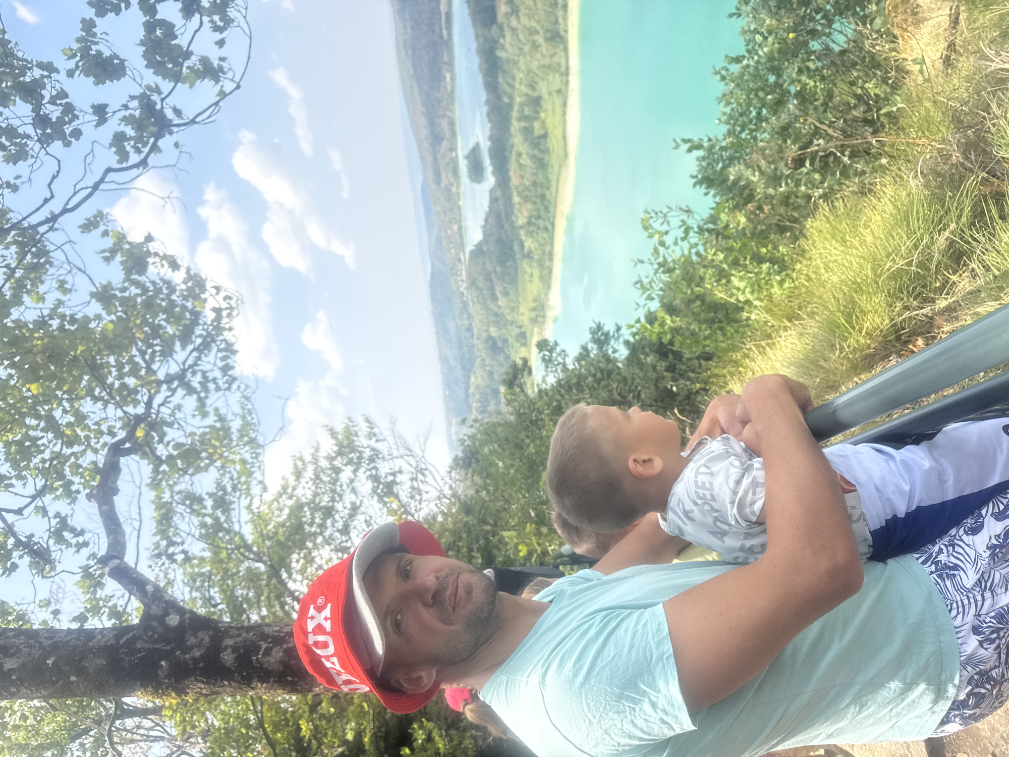 In the city of La Chaux-du-Dombief, France, a man wearing a red bucket hat with "LUX" printed on it and a light blue t-shirt is standing next to a young boy. The man has his arm around the boy. The boy is wearing a light-colored t-shirt with a print and blue and white shorts. They are both looking out at a scenic view of lakes and hills. The environment is outdoors with trees and a bright sky. The image appears to be taken on a sunny day, likely during the daytime.