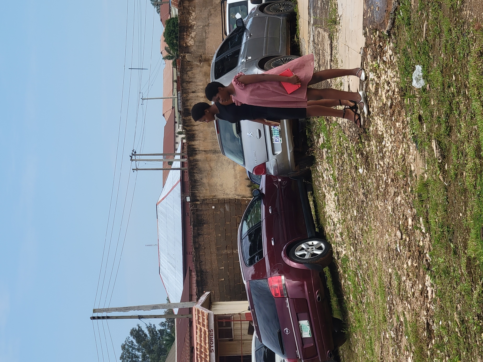Two young females, one wearing a black dress and the other a pink dress, stand in front of parked cars and a building. The female in the black dress holds a book in her right hand. The female in the pink dress holds a red object, possibly a book or tablet, in her right hand. They are standing on a rough, uneven ground with scattered rocks and grass. In the background, there are several cars, including a maroon Opel station wagon and a silver Peugeot. A brick wall runs along the background, with buildings and power lines visible above it. The sky is clear and blue. The scene appears to be outdoors in a relatively undeveloped area, potentially on the outskirts of a city like Gura Top, Nigeria. The presence of parked cars and a building with a sign that reads "STORES" suggests a commercial or residential area.