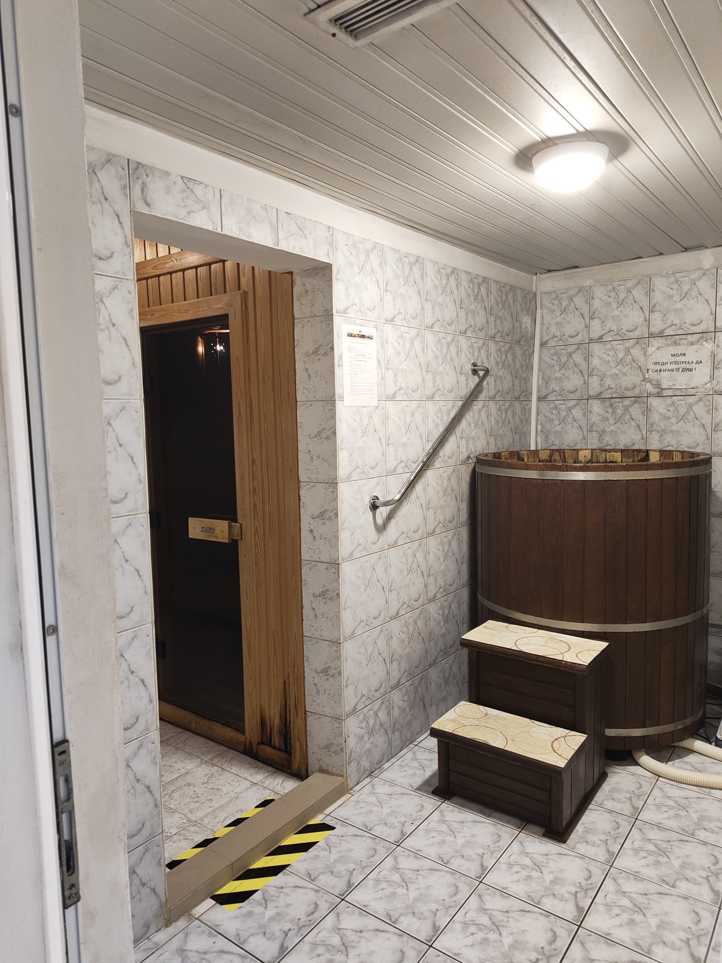 The image shows the interior of a bathroom or spa area in Kozloduy, Bulgaria. The room features tiled walls and floors with a marble pattern. On the right side of the image is a large, wooden barrel tub, which is accessed by two wooden steps. A metal handrail is mounted on the wall next to the tub. A white drainage hose is visible near the base of the tub. On the left side of the image is a doorway leading to a sauna, marked with a yellow and black striped hazard border on the floor. There are two signs mounted on the wall. The lighting is provided by a ceiling-mounted light fixture. The room appears to be well-maintained.