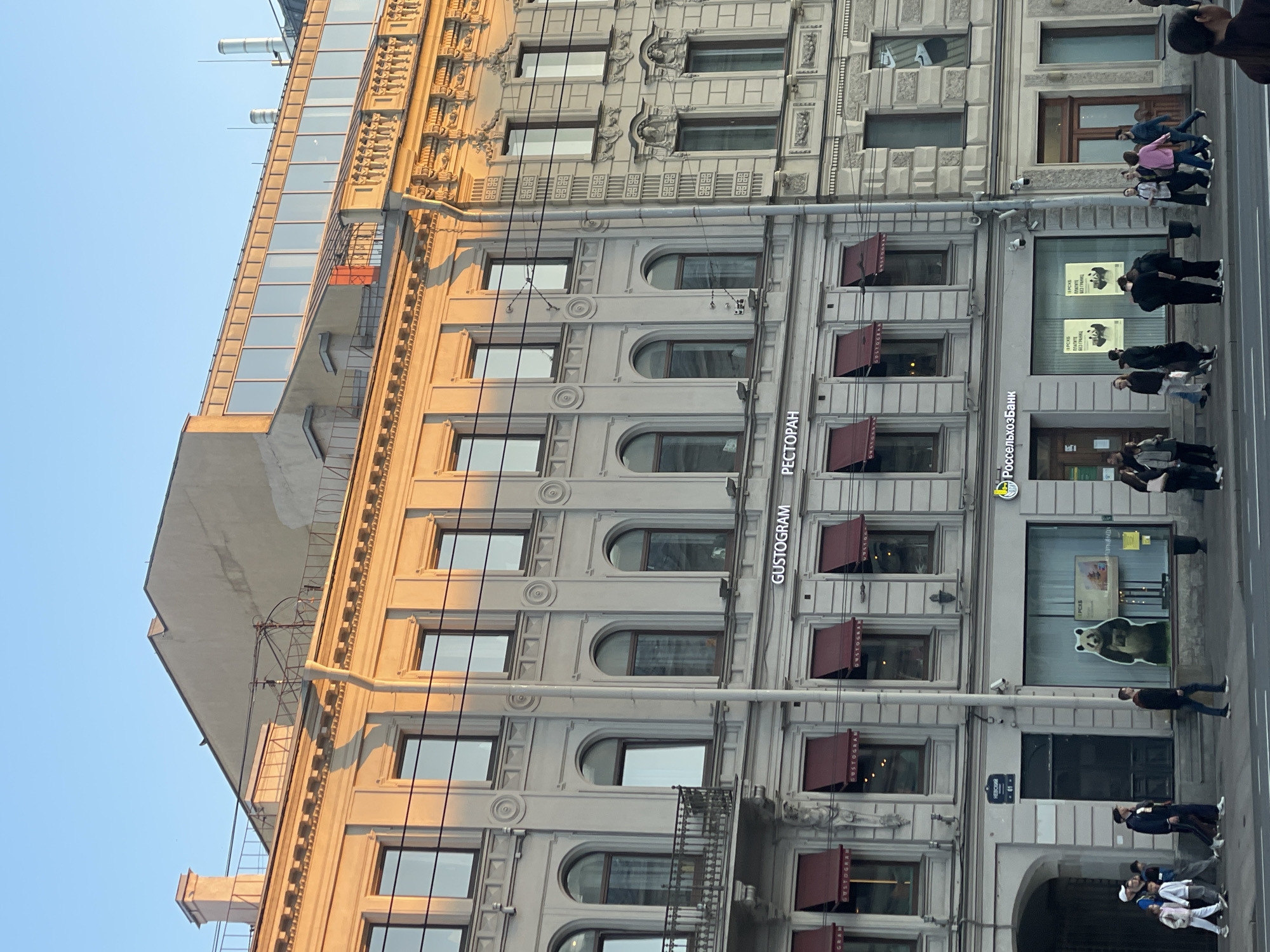 The image shows a multi-story building in what appears to be Saint Petersburg, Russia, as the signs are in Russian. The building is ornate with multiple windows and architectural details. There is a restaurant named "GUSTOGRAM" on the ground floor. People are walking on the sidewalk in front of the building. The photo is taken at an angle. There is a bank named "Россельхозбанк" also on the ground floor of the building. A sign with the number "61-" is visible on the building.