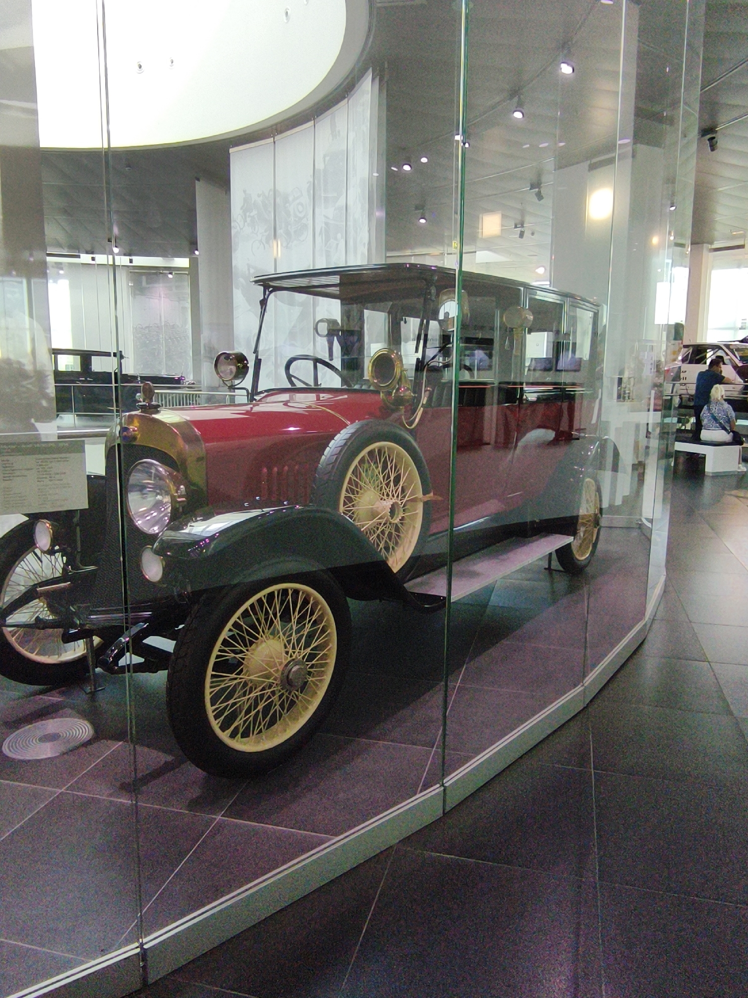 The photograph, taken in the Audi Museum in Ingolstadt, Germany, features a vintage red and dark green automobile displayed behind a glass enclosure. The car, with its prominent spoked yellow wheels and brass accents around the headlights and hood, is positioned on a dark tiled floor. A plaque with text is visible to the left of the car's front wheel. In the background, other exhibits and visitors are faintly visible, with one individual, a person with blonde hair wearing a blue patterned top, seated on a white bench, and another person, appearing to be a male with dark hair, standing near them. The museum environment includes suspended lighting and architectural elements like a curved white ceiling.