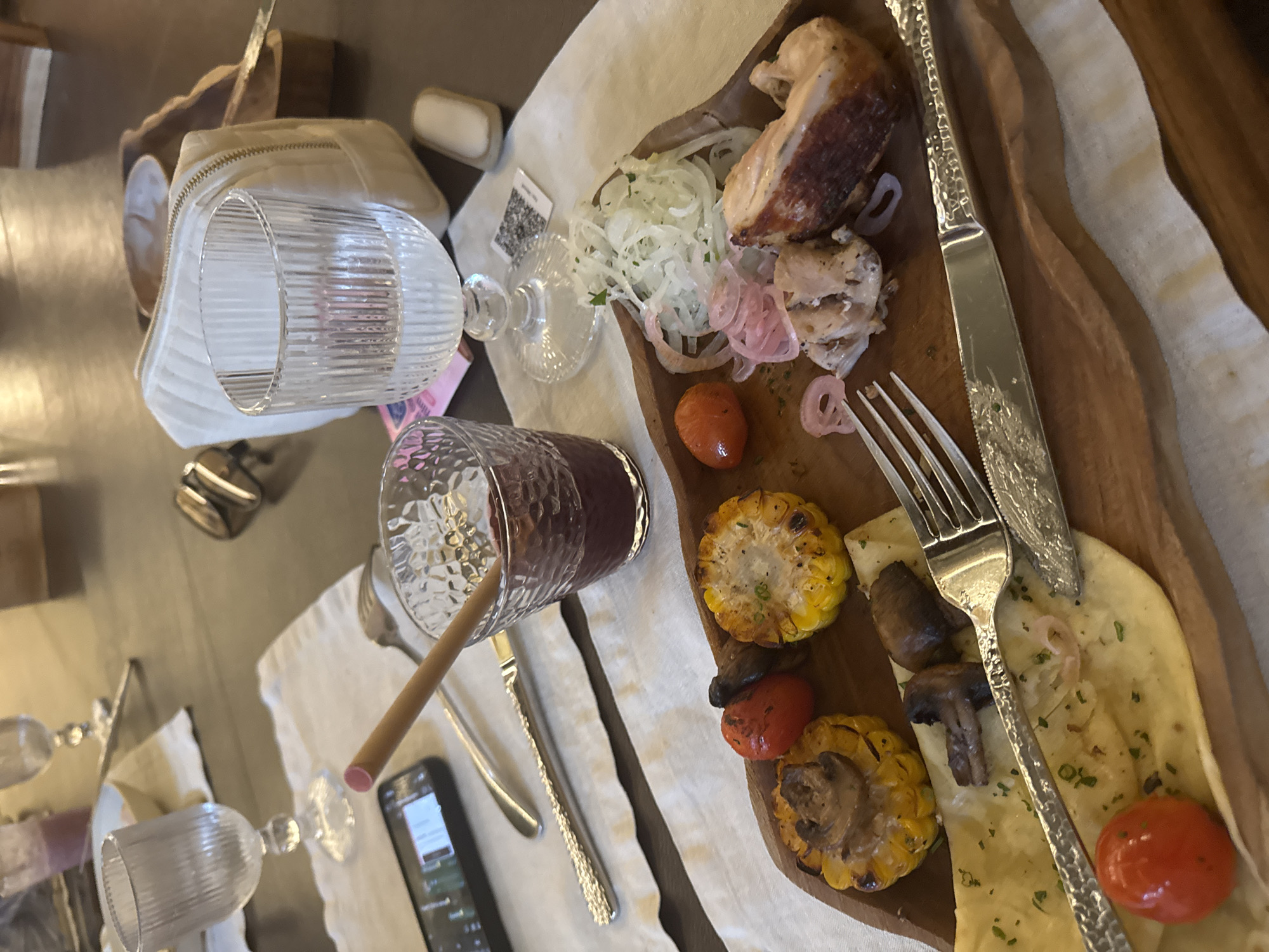 The image shows a table setting in what appears to be a restaurant. A wooden platter holds grilled chicken with onions, grilled corn, tomatoes, mushrooms, and what looks like flatbread. Silverware is placed on the platter and next to a glass of a dark red beverage with a straw. A phone is on the table near the glass. Other glassware, a bag, and personal items are scattered on the table around the platter. The table is covered with a white cloth. Based on the prompt indicating a location in North Kuta, Indonesia, the setting is likely a restaurant there. No people are visible.
