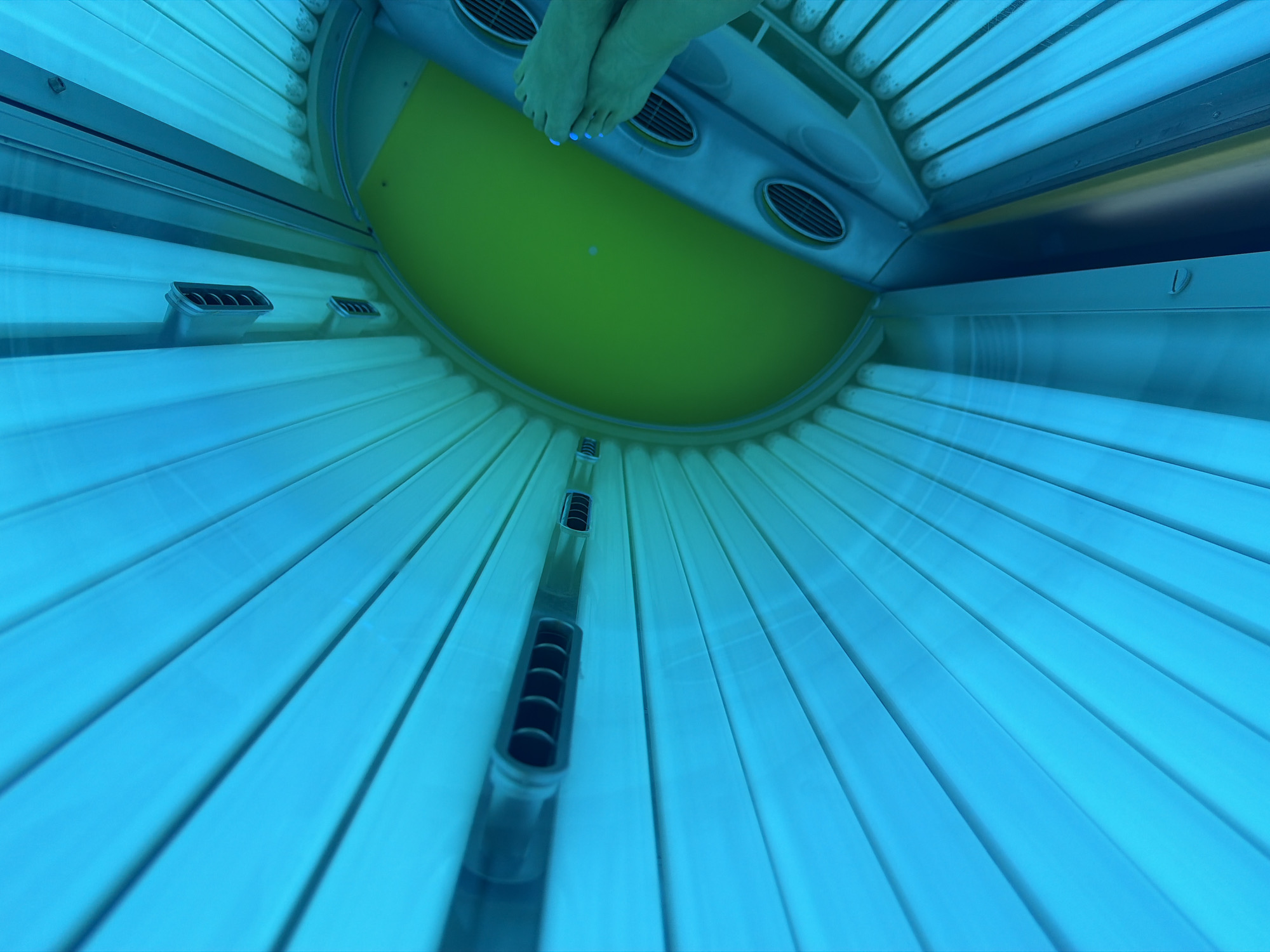 The image shows the interior of a tanning bed, likely in a salon in Kyiv, Ukraine. The bed is illuminated by bright blue UV lights, creating a vibrant and artificial glow. The subject's feet, with light blue painted toenails, are visible at the bottom of the frame, positioned on the tanning bed's surface. The green interior of the tanning bed is also visible, contrasting with the blue lights. The overall environment is enclosed and appears to be designed for personal tanning sessions.