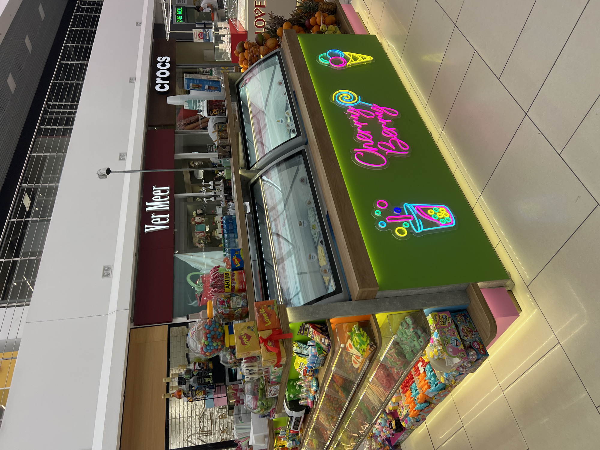 The image captures an ice cream and candy shop located in a modern shopping mall. The shop's facade is brightly lit with neon signs, featuring "Cherry Berry" in pink and yellow, and an ice cream cone and a cup with bubbles in colorful neon. The shop displays a wide variety of colorful candies in clear bins, neatly arranged on tiered shelves. Behind the candy display are two glass-enclosed ice cream freezers. The shop appears to be situated in an indoor public space, likely a mall, with polished tiled flooring and other retail establishments visible in the background, including "Ver Meer" and "Crocs." The overall environment suggests a vibrant and appealing retail setting aimed at attracting customers with its visual presentation of sweets and frozen treats.