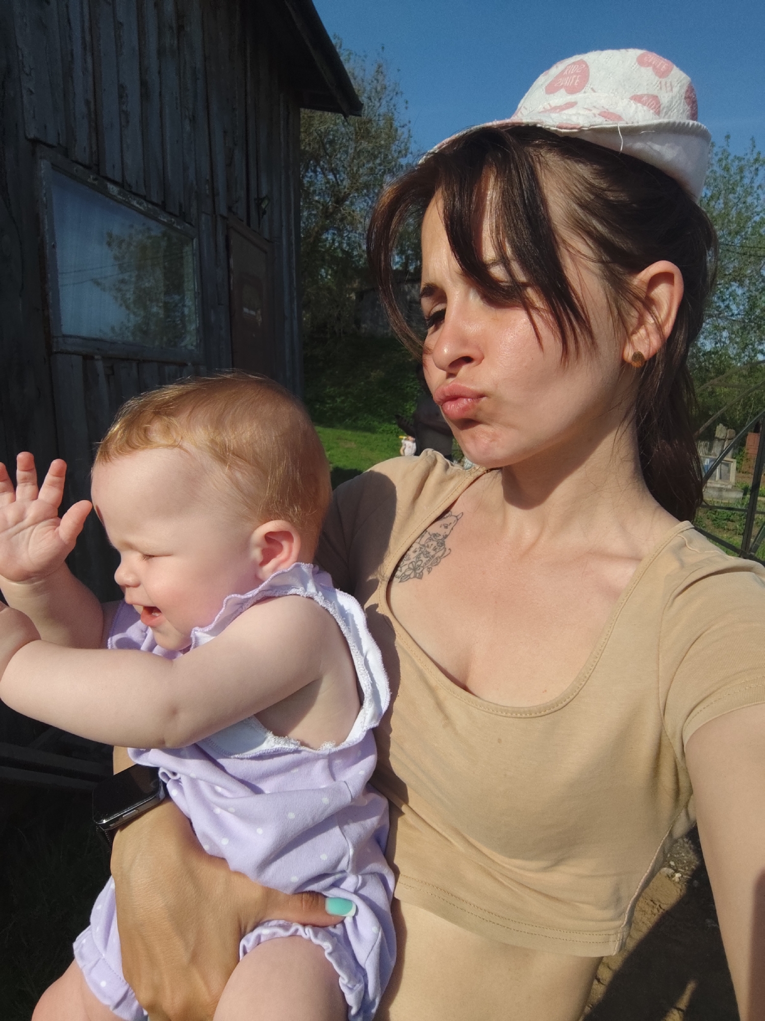 A woman is holding a baby, likely outdoors, in a location that could be Фоки, Russia. The woman has brown hair, fair skin, and a tattoo on her chest. She is wearing a beige top and a hat with pink hearts. The baby has red hair and is wearing a light purple romper with white polka dots. The baby's right hand is raised. The woman is making a kissing face towards the camera. A wooden building is visible in the background with a small window. Trees and a blue sky are also visible.