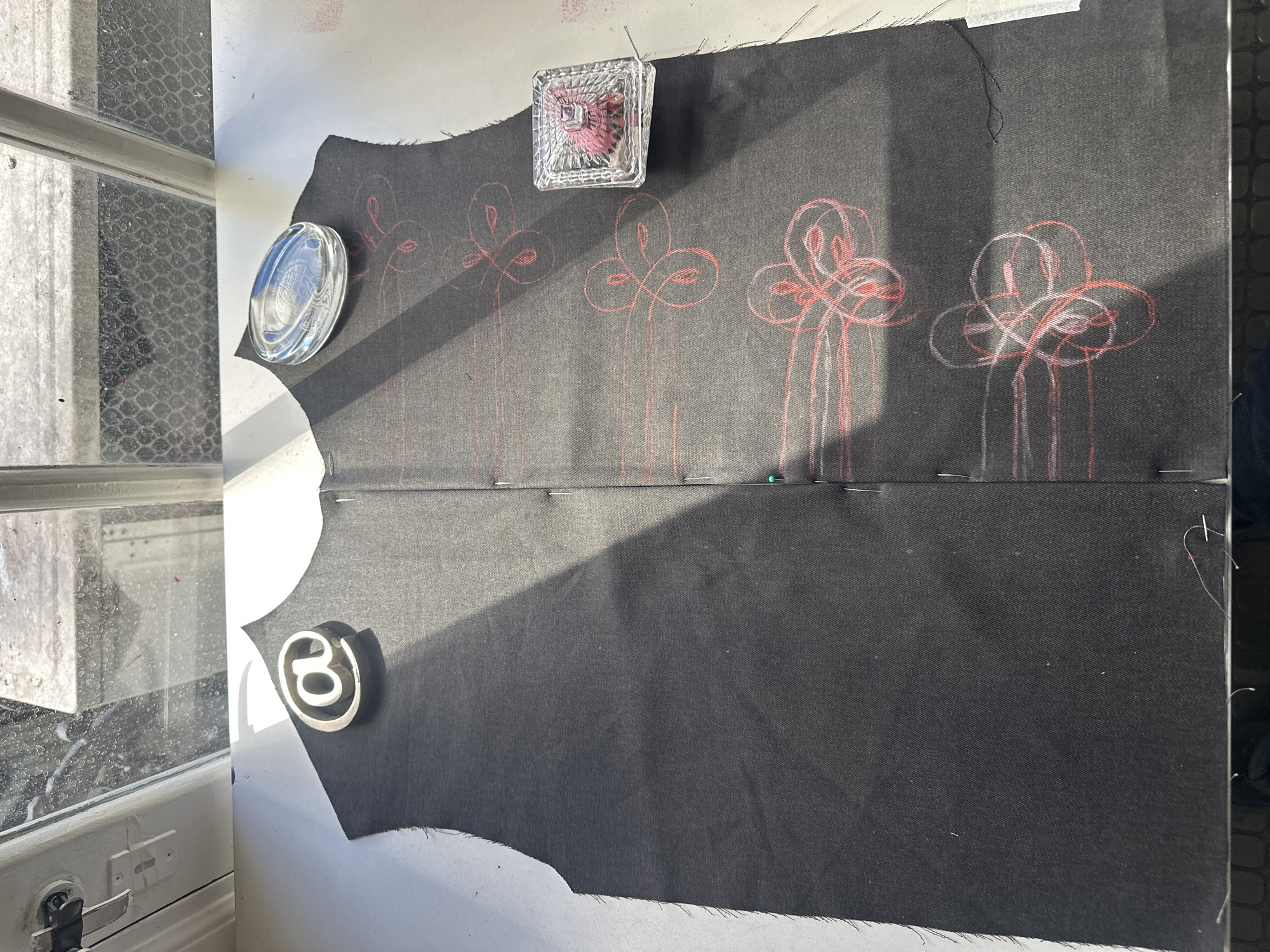 The image shows a piece of black fabric lying on a white surface, possibly a table or desk. The fabric has a series of red, flower-like designs drawn on it, arranged in a row. A clear glass paperweight containing what appears to be red pins rests on the fabric near the top of the frame. To the left, a clear, oval-shaped object and a small, round object with a "3" on it are placed on the white surface near the edge of the fabric. The fabric appears to be pinned together along a horizontal line with multiple silver and one blue-headed pin.
The scene is lit by sunlight, casting shadows across the fabric. A dirty window is partially visible on the left side of the image. The image provides insufficient detail to identify the specific location in Buenos Aires, Argentina, or any other details about people, actions, or interactions in the scene.
