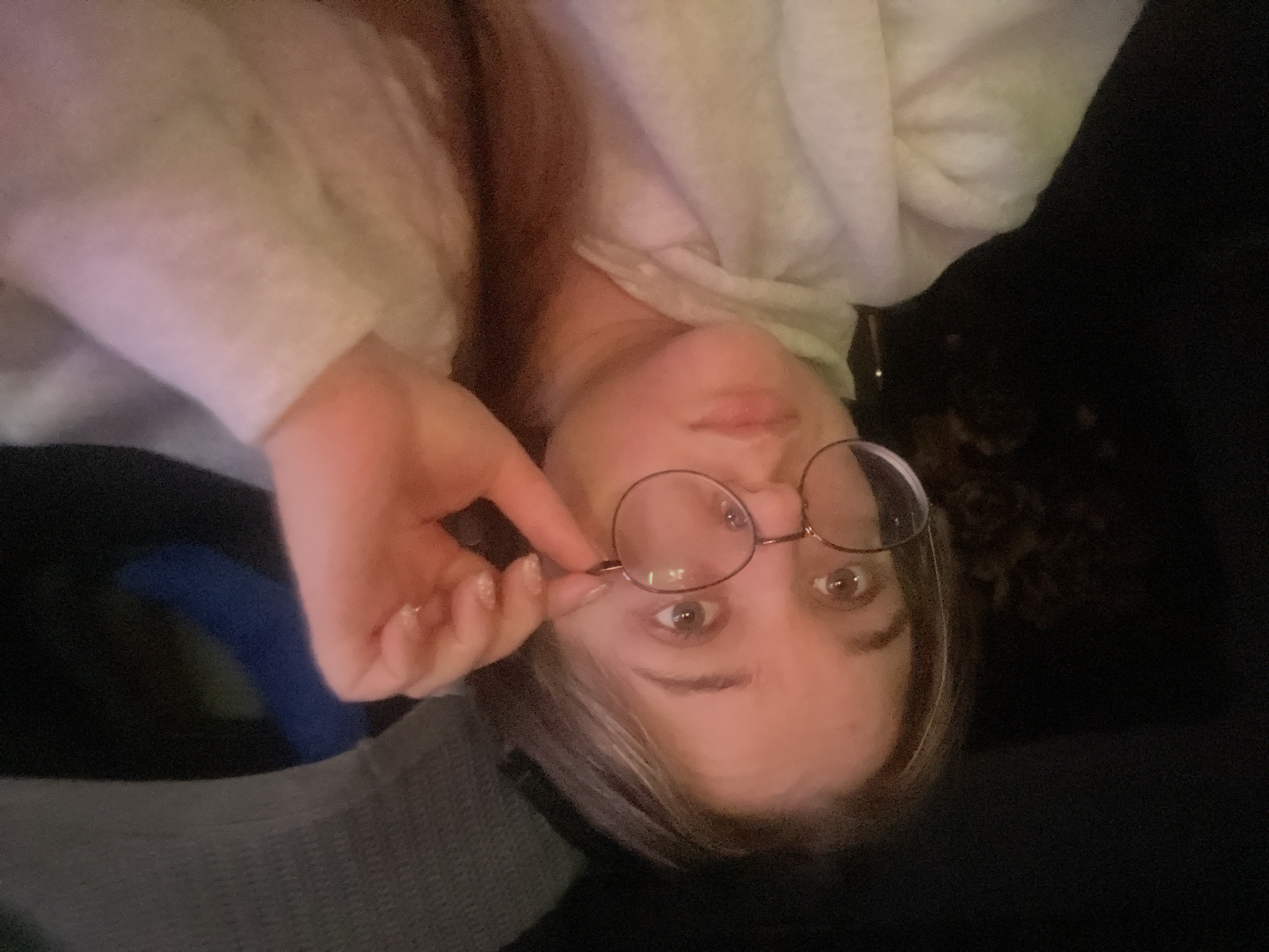 The image shows a close-up, overhead shot of a young person with light brown hair wearing a light-colored hooded sweatshirt and round glasses. Their right hand is raised, holding the bridge of the glasses. The person is looking directly at the camera with their eyes open. The background is dark and out of focus, with a hint of a textured gray object behind the person's head and a blue object to the right. The lighting appears to be artificial, casting a warm glow on the subject's face. The location in the city of Kolomna, Russia, is not discernible from the visual information presented in the image.