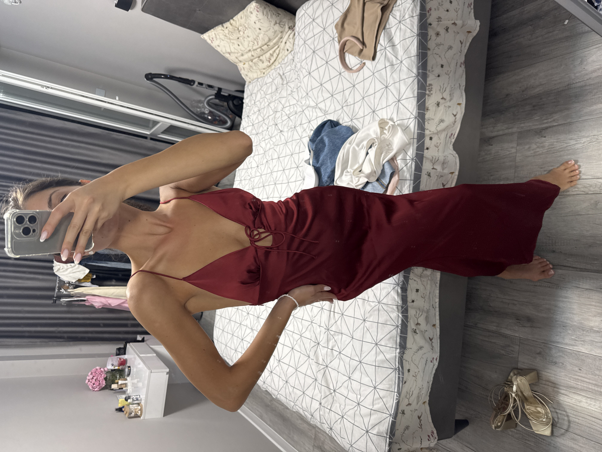 The image depicts a woman standing in a bedroom, reflected in a mirror. She is wearing a dark red, spaghetti-strap slip dress with a tie detail at the bust. Her left arm is bent, with her hand on her hip, and she holds a smartphone in her right hand, obscuring her face. In the background, a bed with a white and grey patterned duvet is visible, with various items of clothing scattered on it, including a blue denim item and a white top. A vanity table with makeup and a vase of pink flowers sits to the left of the frame. To the right, a tall mirror and a vacuum cleaner are present. The floor is wooden. The scene is consistent with a private residence in Burgas, Bulgaria.