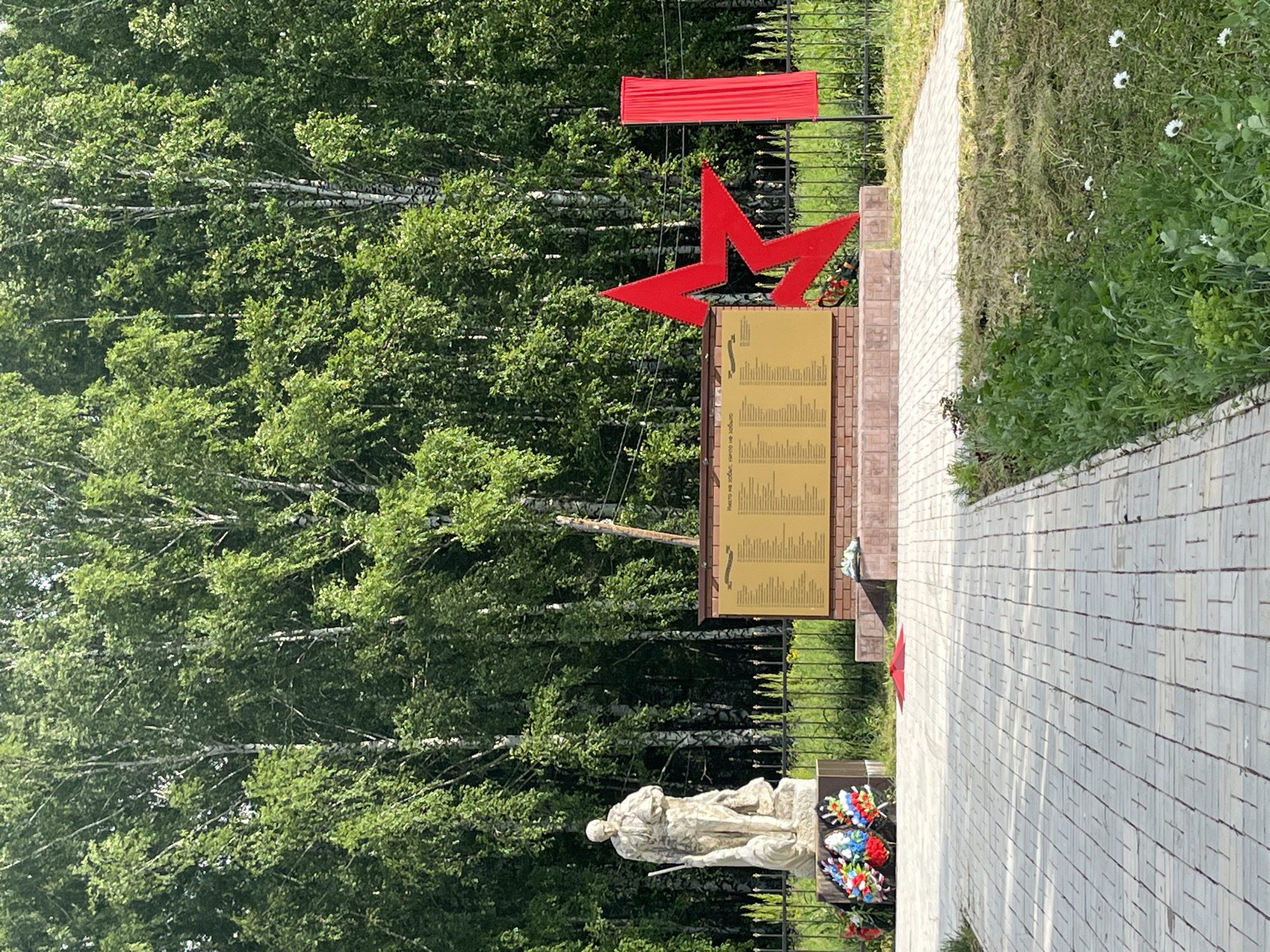 This is a ground-level, eye-level, medium shot of a war memorial located in the city of Bolshoy Gondyr, Russia. The memorial features a stone statue of a soldier holding a helmet and a flag. In front of the statue, there is a wreath of flowers. Behind the statue is a brick wall with a plaque listing names. To the right of the plaque is a large red star and a red banner. The memorial is surrounded by a metal fence. The background consists of a dense forest of birch trees with green leaves. A paved walkway leads to the memorial, and there is grass and some small white flowers in the foreground. The lighting suggests it is daytime with sunlight.