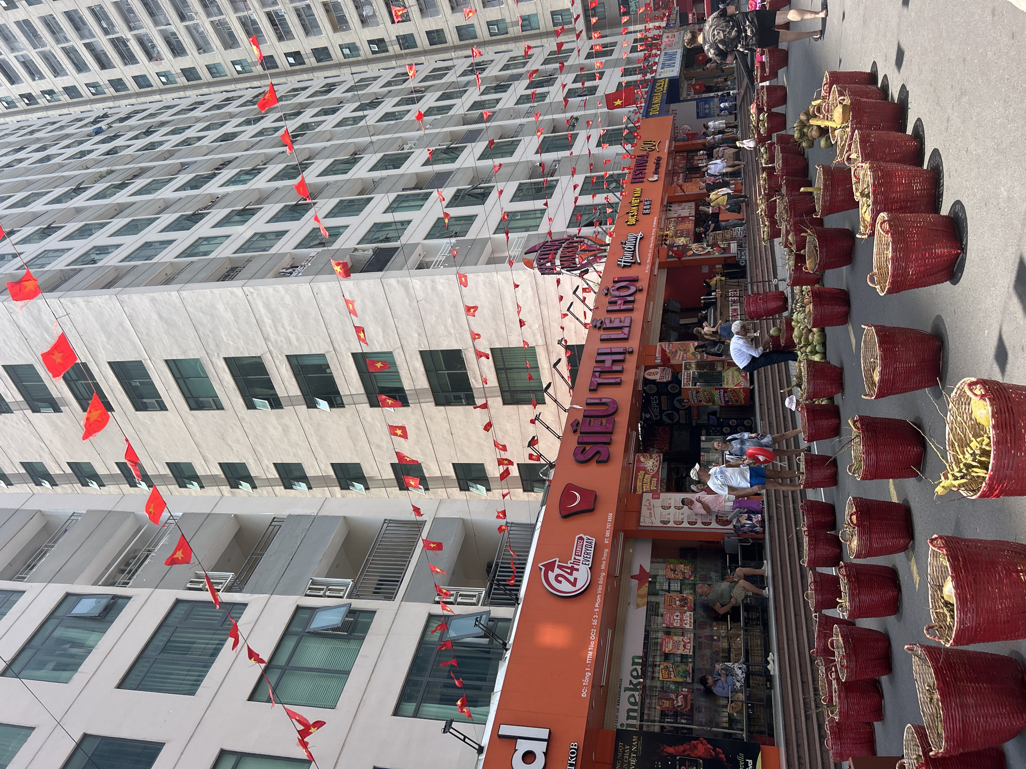 The image depicts an outdoor scene in Nha Trang, Vietnam, specifically near the 'Siêu Thị Lễ Hội' festival supermarket. The supermarket's large orange signage is visible. Many red Vietnamese flags are strung up, decorating the storefront and the tall residential building in the background. Red woven baskets, filled with coconuts, are placed in rows along the street, apparently for sale. Several people are visible near the store, including shoppers and vendors. The image appears to be taken during daylight hours, with bright sunlight illuminating the scene. The text on the supermarket's signage includes details such as "24 hr SERVICE EVERYDAY" and address information related to Pham Van Dong Street.