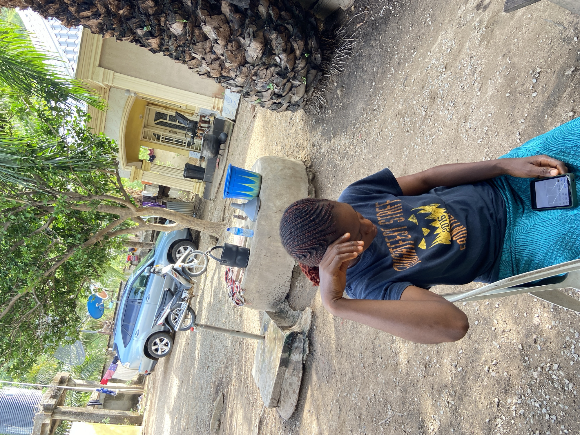 A woman with braided hair is sitting on a white chair outside a house in Mararaba, Nigeria. She is wearing a dark t-shirt and a blue patterned wrap. Her left hand is resting on her forehead, and she is holding a smartphone in her right hand. In the background, there is a blue car, a motorcycle, a tree, and a house with a yellow entrance. A blue bucket sits on a concrete structure next to plastic water bottles. The ground is dirt with small pebbles. The trunk of a palm tree is visible in the upper part of the image.