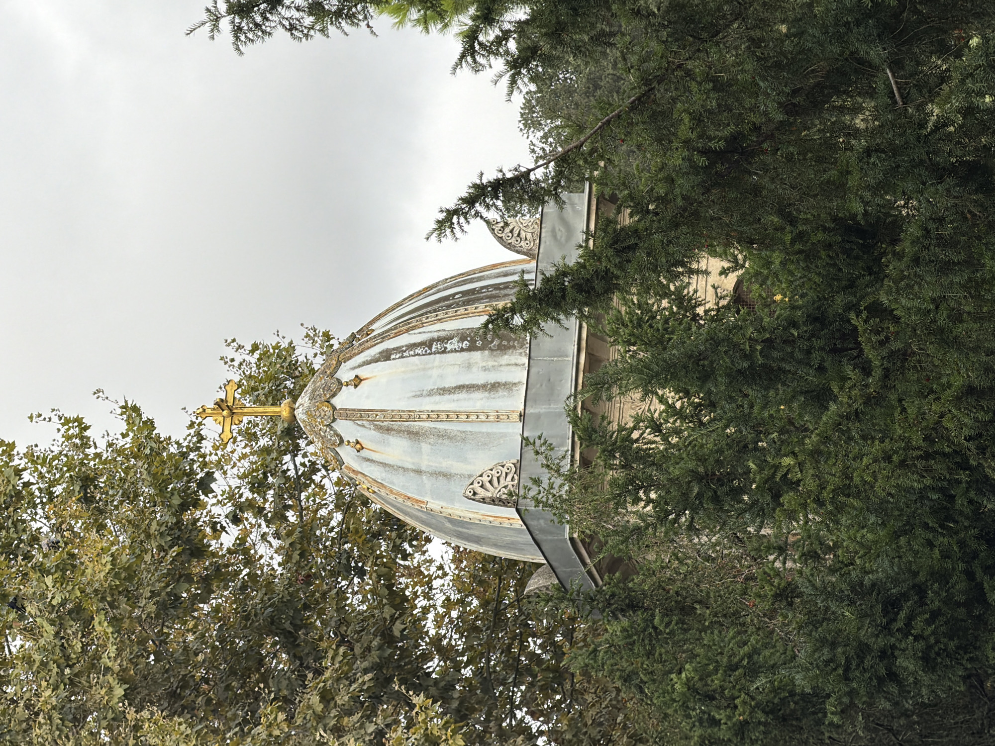 A religious structure's dome and golden cross are visible through dense evergreen trees. The dome is weathered and appears to be made of metal, with ornate detailing along its base and top. The golden cross is positioned at the apex of the dome. Trees with green foliage surround the structure, suggesting an outdoor setting. The sky is overcast, casting a diffused light on the scene. This structure is the dome of the Russian Orthodox Church of St. Panteleimon, located in the Kuzguncuk neighborhood of Üsküdar, Istanbul, Turkey.