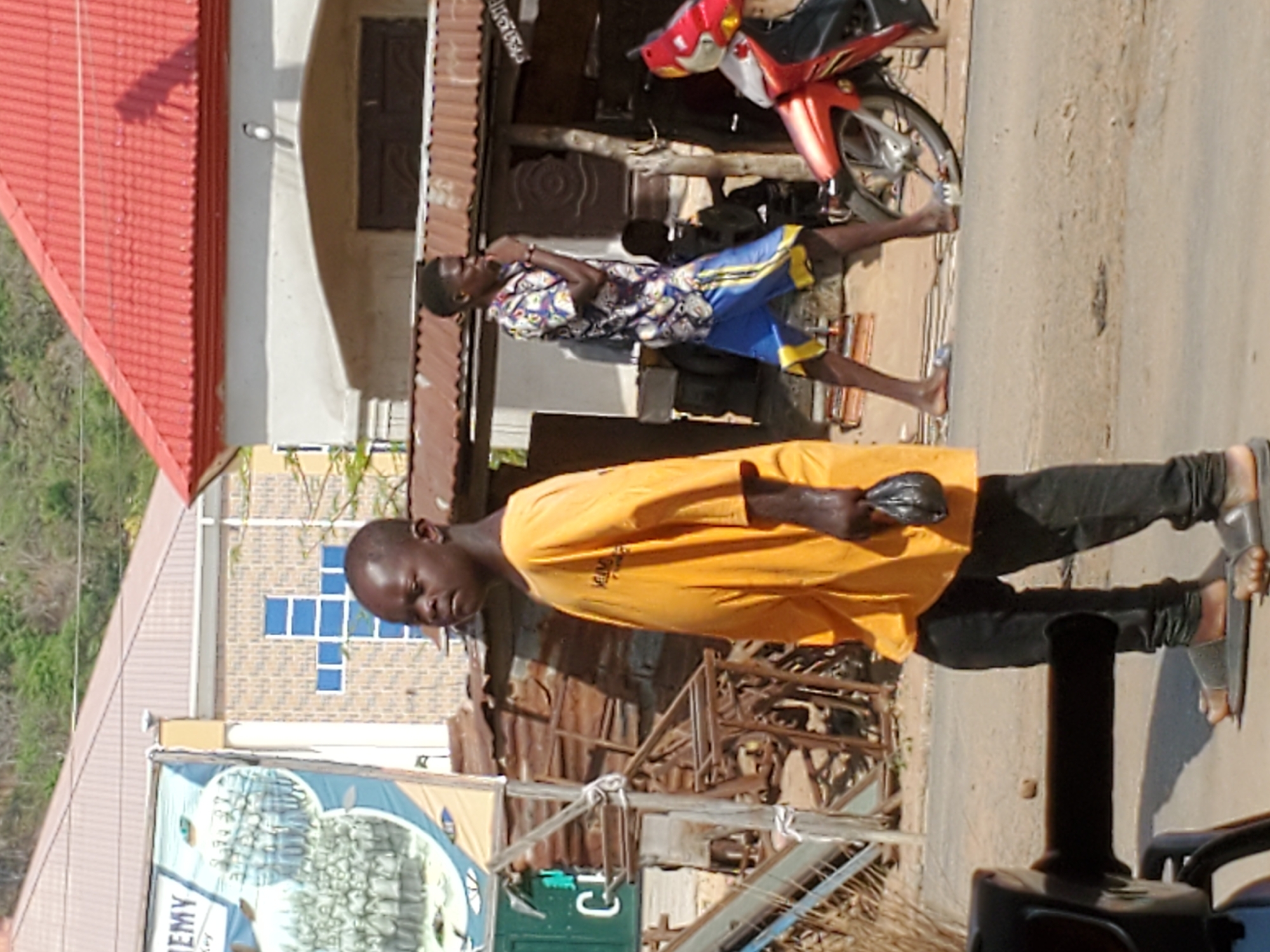 The image shows two African men walking on a street in Mararaba, Nigeria. The man in the foreground wears an orange t-shirt and black pants, and appears to be holding something in his left hand. He is facing the left side of the image. He is wearing sandals.
The second man walks behind him, wearing a patterned, short-sleeved shirt and blue shorts with yellow trim. He appears to be barefoot.
Behind them, a parked motorcycle is visible. Buildings line the street, including one with a red roof and a sign for "Academy". The buildings appear to be constructed from brick and corrugated metal. The environment appears urban and potentially impoverished.