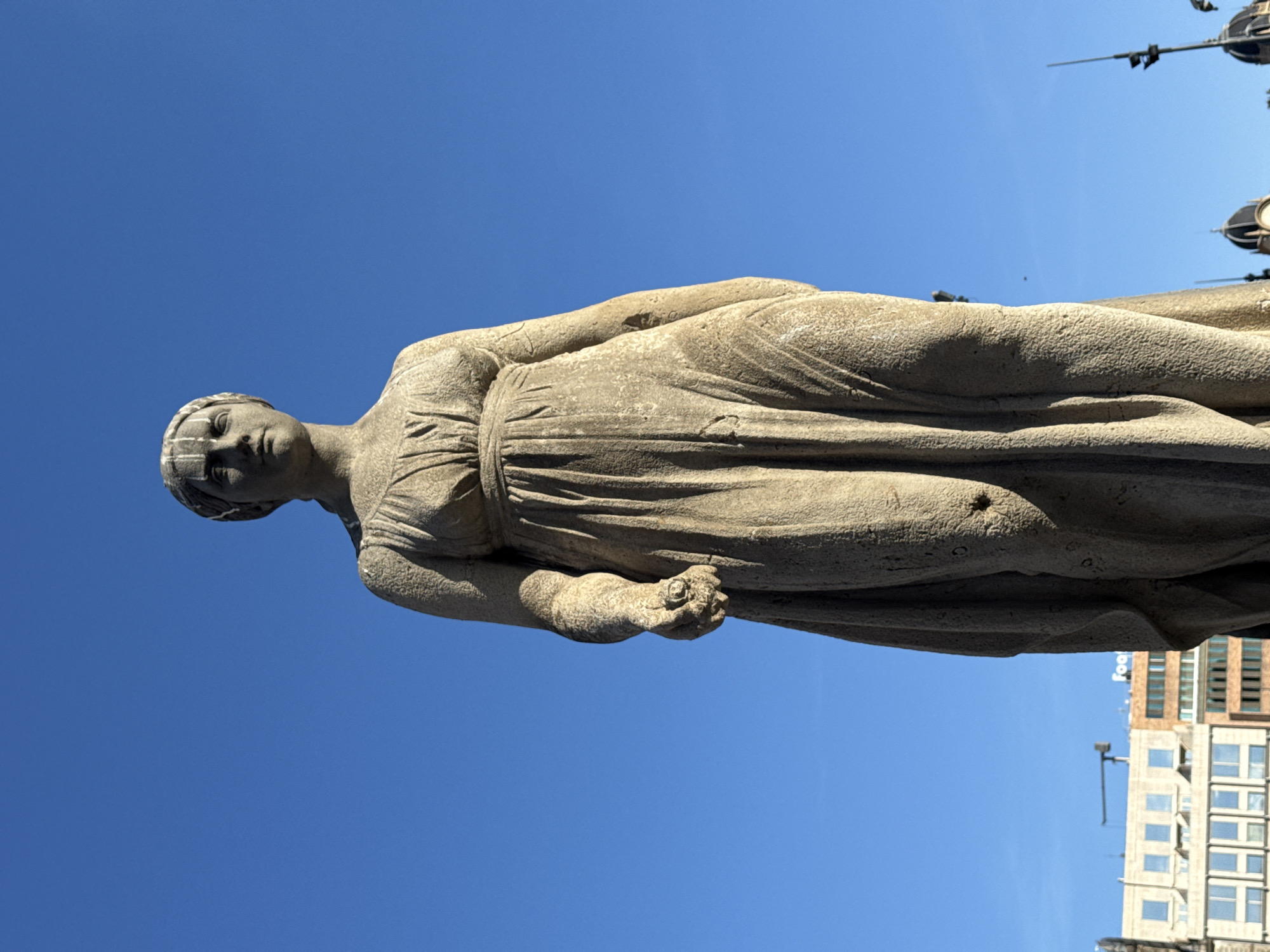 A stone statue of a woman is depicted against a clear blue sky. The statue is weathered and appears to be of classical style, draped in a toga-like garment. She holds something in her right hand. Modern buildings and a decorative lamp post are visible in the background, indicating the statue's location within a city. Given the architectural style of the buildings, this is likely in Barcelona, Spain, specifically at Plaça de Catalunya (Catalunya Square).