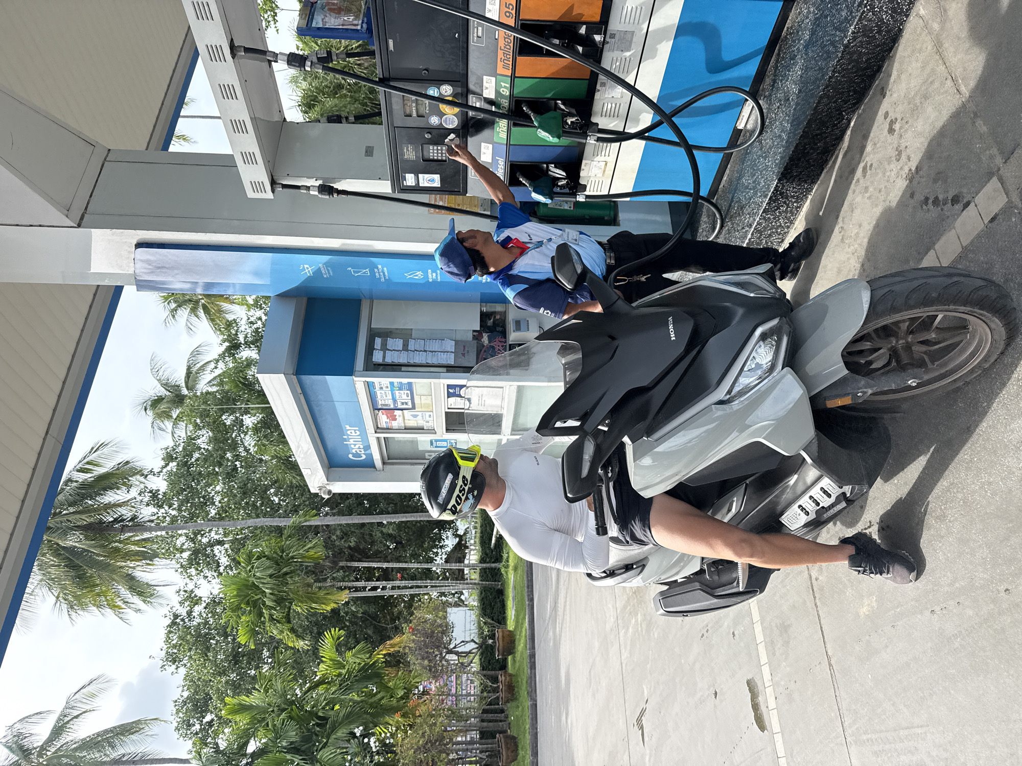A man on a scooter is at a gas station in Ko Samui District, Thailand. He is wearing a white long-sleeved shirt, black shorts, a helmet, and yellow goggles. A gas station attendant in a blue and red uniform and a blue cap is at the pumps, interacting with the control panel. The gas pumps have labels in Thai script. Palm trees and lush green foliage are visible in the background. The scene is illuminated by daylight.