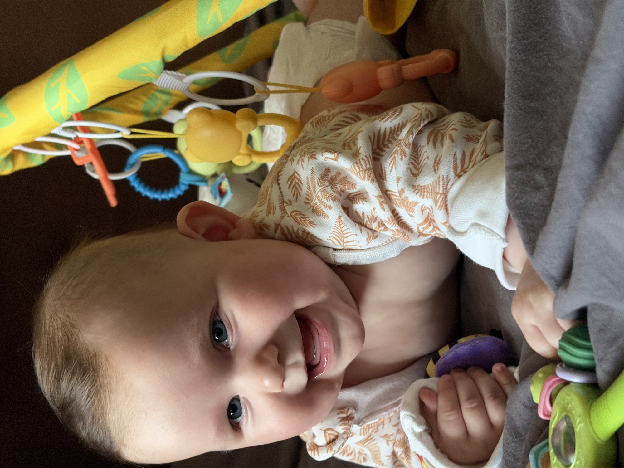 A close-up, portrait-oriented photograph captures a happy baby lying on its back, engaged in play. The baby, with light blonde hair and bright blue eyes, looks directly at the viewer with a wide, joyful smile, its tongue playfully peeking out. It wears a white long-sleeved garment adorned with a delicate brown and orange fern pattern.
Above the baby is a vibrant yellow play gym arch, decorated with green leaf motifs. Several colorful toys hang from the arch, including a yellow animal-shaped teether, an orange ring, and a blue textured ring, indicating a stimulating environment for the infant. The baby is actively interacting with handheld toys, gripping a purple and yellow soft toy in its right hand and a green, multi-textured rattle in its left.
A soft, gray blanket or fabric is visible along the right side of the frame, suggesting a comfortable indoor setting. The lighting is consistent and even, typical of an interior space during the day. The scene exudes an atmosphere of innocence and contentment. No specific time of day or weather conditions can be inferred, nor are there any discernible location markers within the image. No visible text is present. The provided location context for this scene is Chaikovsky, Russia.