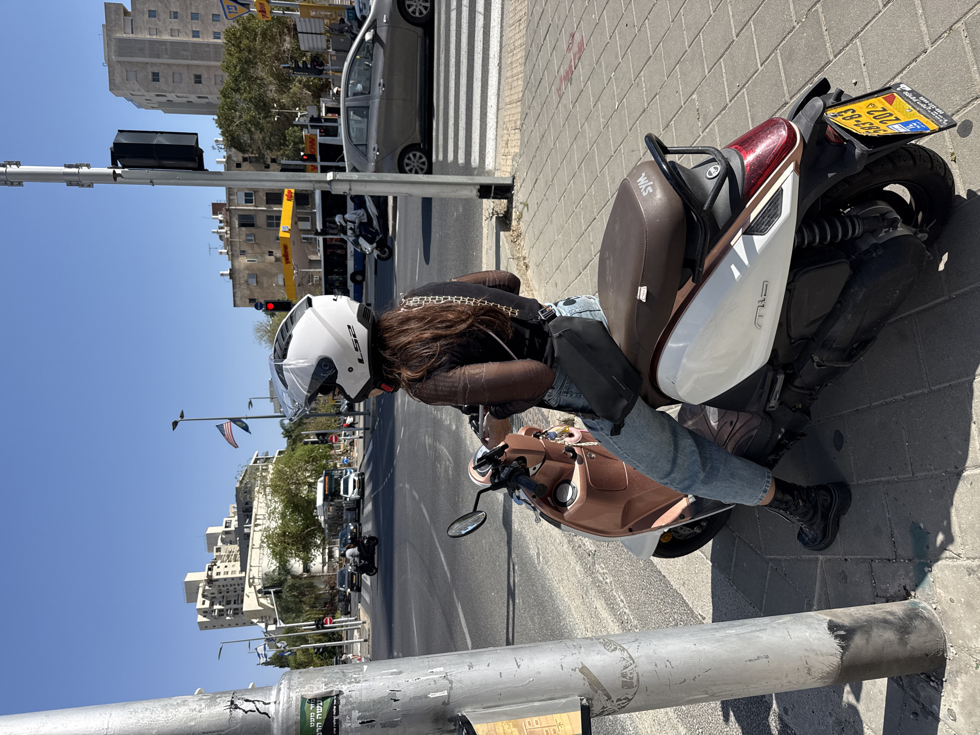 A woman wearing a white LS2 helmet, a dark long-sleeved top with a chain-like pattern, blue jeans, and black combat boots, is seated on a rose gold and white SYM scooter. She has long, dark brown hair flowing from beneath her helmet and carries a black shoulder bag. She appears to be paused on the side of an urban street, possibly waiting at a red traffic light, as one is visible.
The scene depicts a sunny day with a clear blue sky, casting strong shadows on the paved sidewalk and street. In the background, several multi-story residential or commercial buildings line the street. Other vehicles, including cars and scooters, are visible on the road and parked. A white crosswalk is painted across the street in the distance. Street poles, traffic lights, and some greenery from trees are also part of the urban landscape. An Israeli flag and another flag resembling the US flag are seen flying from poles in the distance.
Notable text includes the brand "LS2" on the woman's helmet and "SYM" and "M/S" on the scooter's brown seat. The scooter's rear license plate is yellow with blue text, indicating an Israeli plate, and shows "ישראל" (Israel) along with the number "202-31-83". Fragments of Hebrew text, possibly from an advertisement or public message, are visible on a sticker attached to a street pole. "Yellow pages" (דפי זהב) signs are visible on some buildings. The setting is consistent with the provided location context of Ramat Gan, Israel.