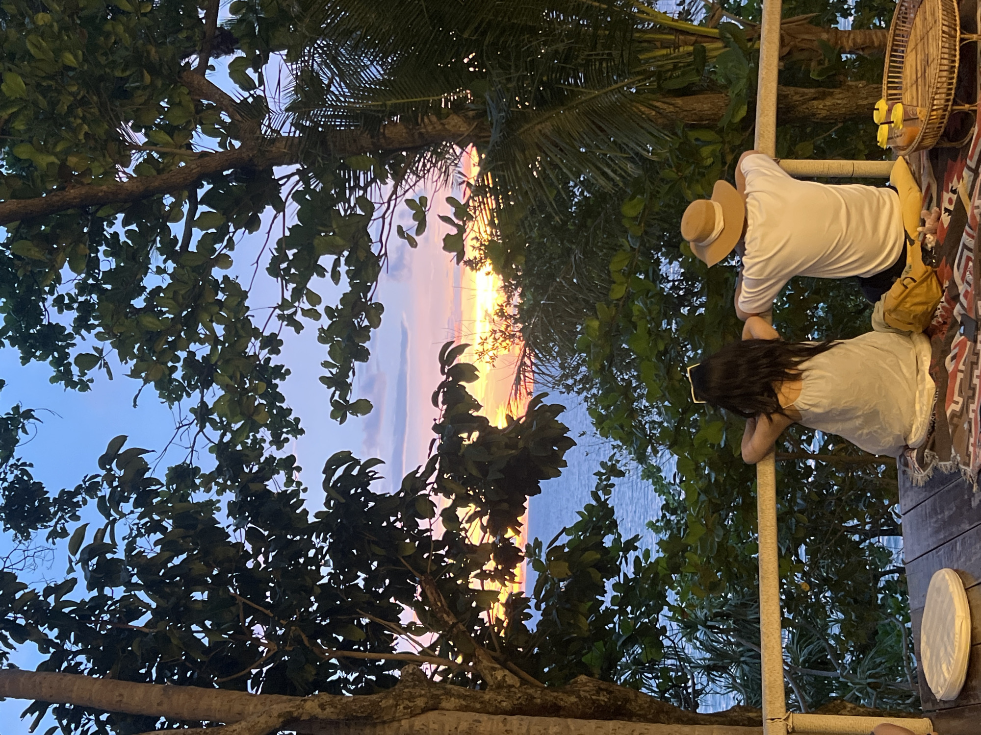 Two people, a woman and a man, are seated on a wooden deck overlooking the ocean at sunset. The woman is on the left, wearing a light-colored dress and sunglasses on her head. The man is on the right, wearing a white t-shirt and a wide-brimmed hat. They are both leaning on a railing, facing the sunset over the water. In the foreground, there is a colorful rug, a yellow backpack, and a woven tray with two cups of orange liquid. Lush green trees frame the scene, with a large tree trunk on the left and palm fronds on the right. The sky is painted with hues of blue, pink, and orange as the sun dips below the horizon, casting a warm glow on the ocean. This serene setting is likely a viewpoint in Karon, Thailand, offering a picturesque view of the Andaman Sea.