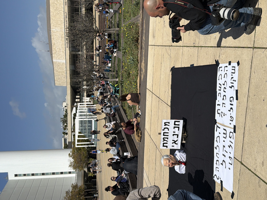 An outdoor scene is captured at Habima Square in Tel Aviv-Yafo, Israel, on a sunny day. The foreground features a protest or artistic display centered on a black fabric spread across light-colored paving stones. Emerging from the black fabric is a life-sized bust of a male figure resembling Benjamin Netanyahu, wearing a white shirt and red tie, with its right index finger pressed against its lips in a "shushing" gesture. Adjacent to the bust, a white sign on a stand displays the Hebrew text "מלחמת נתניהו" ("Netanyahu's War"). Two additional white signs are taped flat on the pavement below the black fabric, each with identical Hebrew text: "הכל כשר ? להכריח שיקול פוליטי ?" ("Is everything permissible? To force a political consideration?").
To the right of this display, a male photographer with a shaved head and beard is crouched, aiming a professional Canon DSLR camera with a large lens at the display. Another lens is visible hanging from a strap around his neck. Partially visible on the far left, a person's leg and foot stand near the display.
In the midground, numerous individuals are seated on wooden tiered benches and scattered throughout a landscaped park area with bare-branched trees and some yellow flowers. Many are casually dressed, conversing, or relaxing, some on the grass or in a green recliner.
The background features prominent architectural structures. On the far left stands a tall, white, multi-story building with a distinct cylindrical element, part of the Habima National Theatre complex. To the right, a large, rectangular, beige building is identified as the "CHARLES BRONFMAN AUDITORIUM" by text visible on its facade. The sky above is clear blue with sparse white clouds. Pedestrians are observed walking across the paved square in the distance.