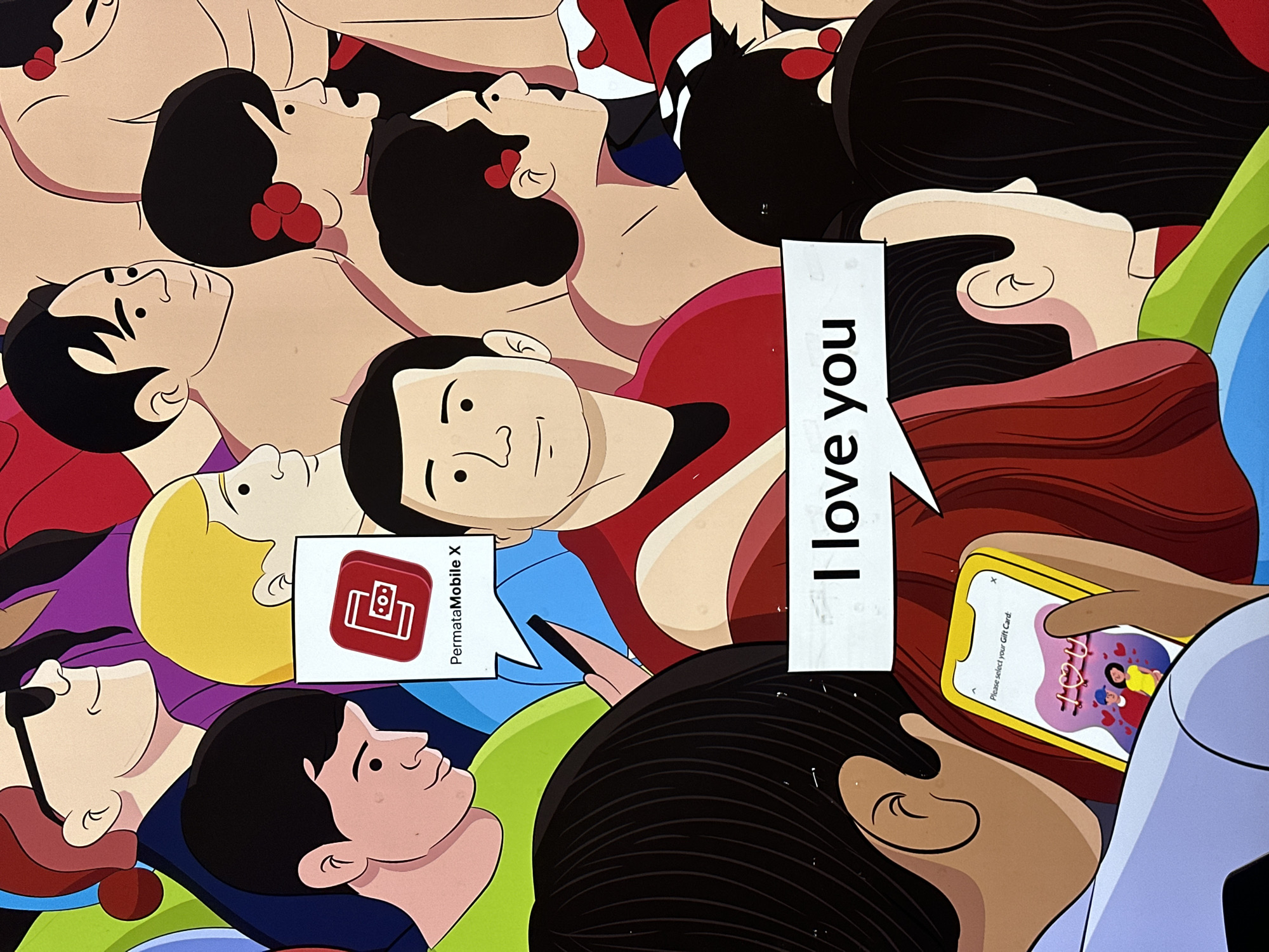 The image is a crowded composition of cartoon-style people lying close together. A speech bubble above one person says "I love you". Another speech bubble contains the text "PermataMobile X" above a red icon. A person in the lower right is holding a smartphone with the text "I love you" and a picture of two cartoon people on the screen. There are no other visual cues that suggest the image is from North Kuta, Indonesia.