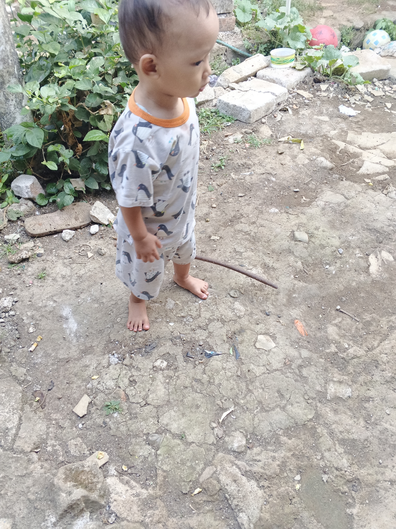In Warungkiara, Indonesia, a young child stands barefoot on a dusty, uneven ground composed of packed dirt and scattered rocks and debris. The child is wearing a light-colored romper with a repeating pattern of penguins and an orange trim around the neckline. The child is facing to the right, with their head turned slightly to observe something out of frame. To the left of the child, a dense green bush with broad leaves grows near a rough, vertical surface, possibly a tree trunk or wall. The background shows more of the cluttered outdoor environment, with more rocks, discarded materials, and some green vegetation. The overall scene depicts an outdoor play area or yard in a residential setting.