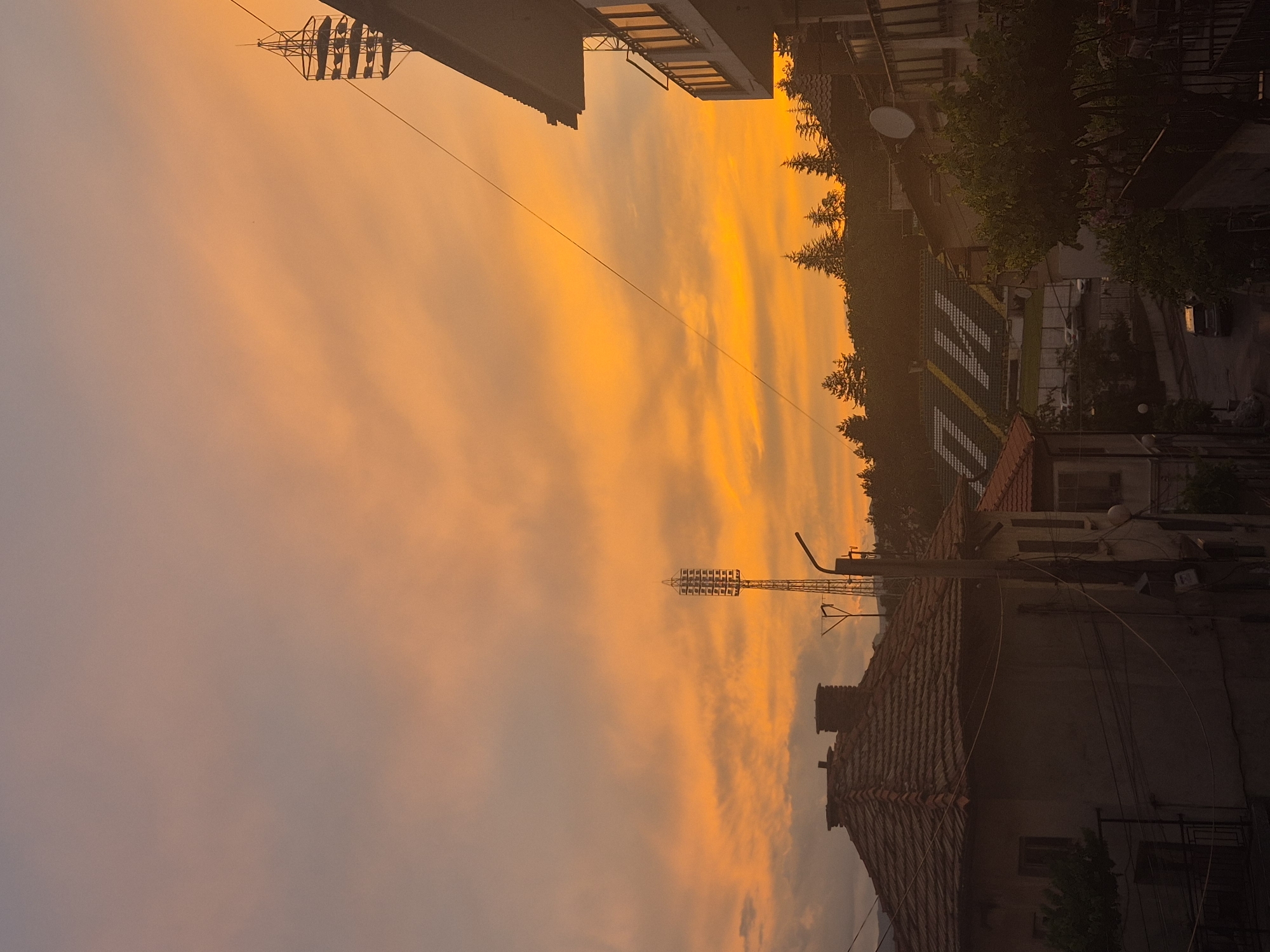 The image depicts a sunset over a town, likely in an unspecified location. The sky is filled with vibrant hues of orange and yellow, with some clouds interspersed. In the foreground, the rooftops of several buildings are visible, some with tiled roofs. A tall radio tower stands prominently in the middle ground, along with a lamppost and electrical wires. In the background, a stadium with seating is discernible, surrounded by trees. The overall impression is of a residential area at dusk, with the natural beauty of the sunset contrasting with the urban landscape. There are no people visible in the image.