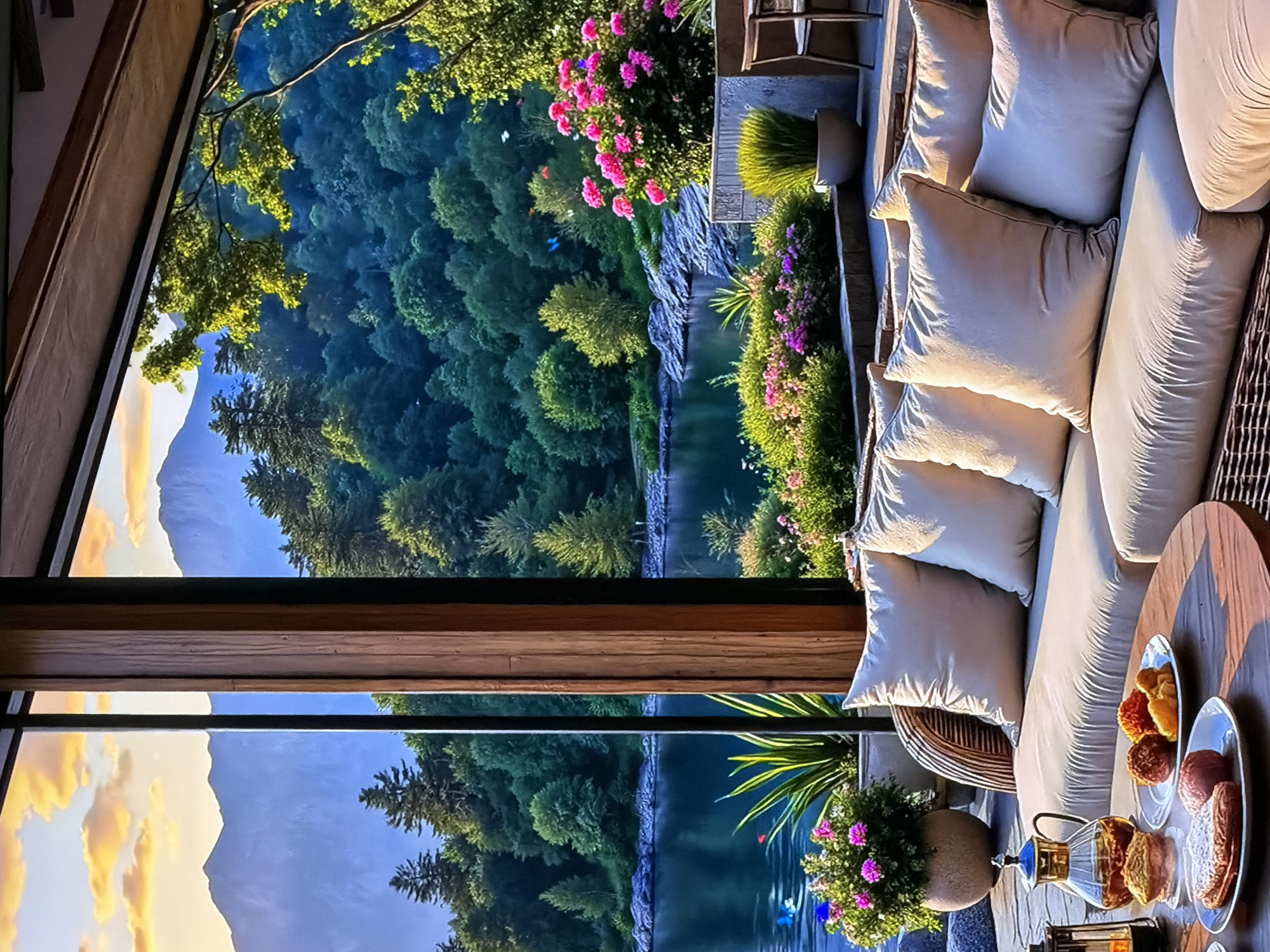 An outdoor setting, possibly a patio or balcony, overlooks a serene natural landscape. In the foreground to the right, a comfortable L-shaped outdoor sofa with numerous light-colored cushions suggests an inviting relaxation spot. A round wooden table in front of the sofa holds a clear glass teapot with a golden top, several glasses, and white plates laden with various pastries or desserts, some dusted with powdered sugar, others appearing caramelized or nut-covered. Potted plants with vibrant pink and purple flowers, alongside pots of tall green grass, decorate the seating area.
Beyond the patio, the scene transitions to a dense forest of varying shades of green, with a calm body of water, likely a river or lake, winding through the midground. Rocky outcrops line the water's edge. Further in the background, visible through a large window or opening on the left, majestic mountains rise under a sky painted with warm, golden-hued clouds, indicating either sunrise or sunset.
The soft, warm light illuminates the scene, highlighting the textures of the cushions and the details of the food, and casting a pleasant glow on the natural elements. The overall atmosphere is one of tranquility and luxurious comfort, designed for enjoying refreshments amidst a breathtaking natural vista. No people are visible, and there is no visible text.