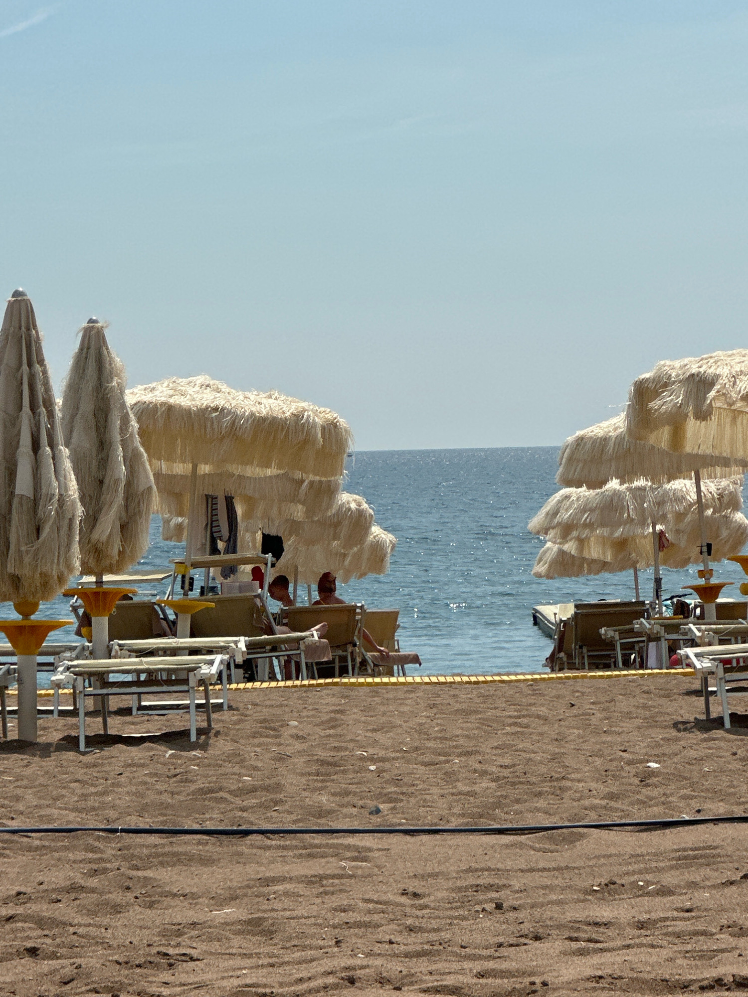 This is a daytime, eye-level shot of a beach scene in Boreti, Montenegro. The foreground is dominated by dark brown sand with some small pebbles. A black cable runs horizontally across the sand. In the middle ground, several beach chairs and umbrellas are set up. The umbrellas have a thatched, straw-like appearance and are positioned over lounge chairs. Some people are visible sitting in the chairs, facing the water. The sea is a calm, blue expanse in the background, with a faint horizon line under a pale blue sky. The water has a slight shimmer, suggesting sunlight. There is a small pier or platform visible extending into the water. The overall impression is a relaxed, sunny beach day.
