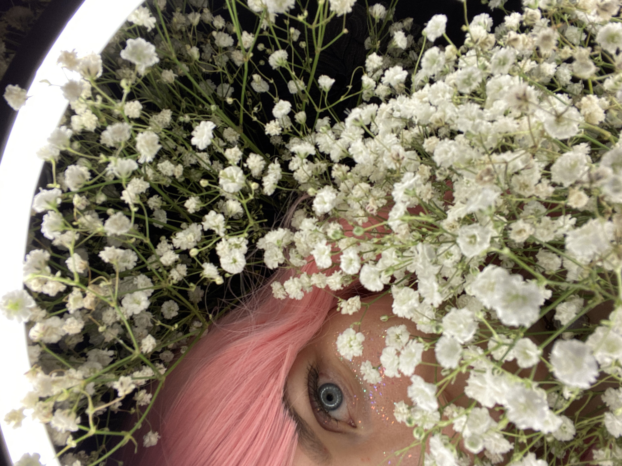 This close-up image features a segment of a person's face, framed by an abundance of small white flowers, resembling Baby's Breath. The visible subject includes a striking blue eye with a dark pupil and natural eyelashes, adorned with sparkling glitter on the skin around it. A wave of bright pink hair is visible to the left and below the eye, extending under the flowers.
The white flowers are densely packed, creating a soft, voluminous frame around the central features. They are illuminated by a bright, circular white light source, likely a ring light, partially visible on the upper left edge of the frame. The background behind the flowers and person is dark and indistinct, suggesting an indoor setting or a scene deliberately isolated by lighting.
The image portrays a creative or artistic portrait, possibly a beauty shot or a themed photographic session. The careful arrangement of flowers, the vibrant hair color, and the glitter around the eye contribute to a whimsical and ethereal aesthetic. The lighting suggests a focused setup, common for professional or artistic photography. The overall mood is serene and enchanting. This media file was captured in Chaikovsky, Russia. No text is visible in the image.