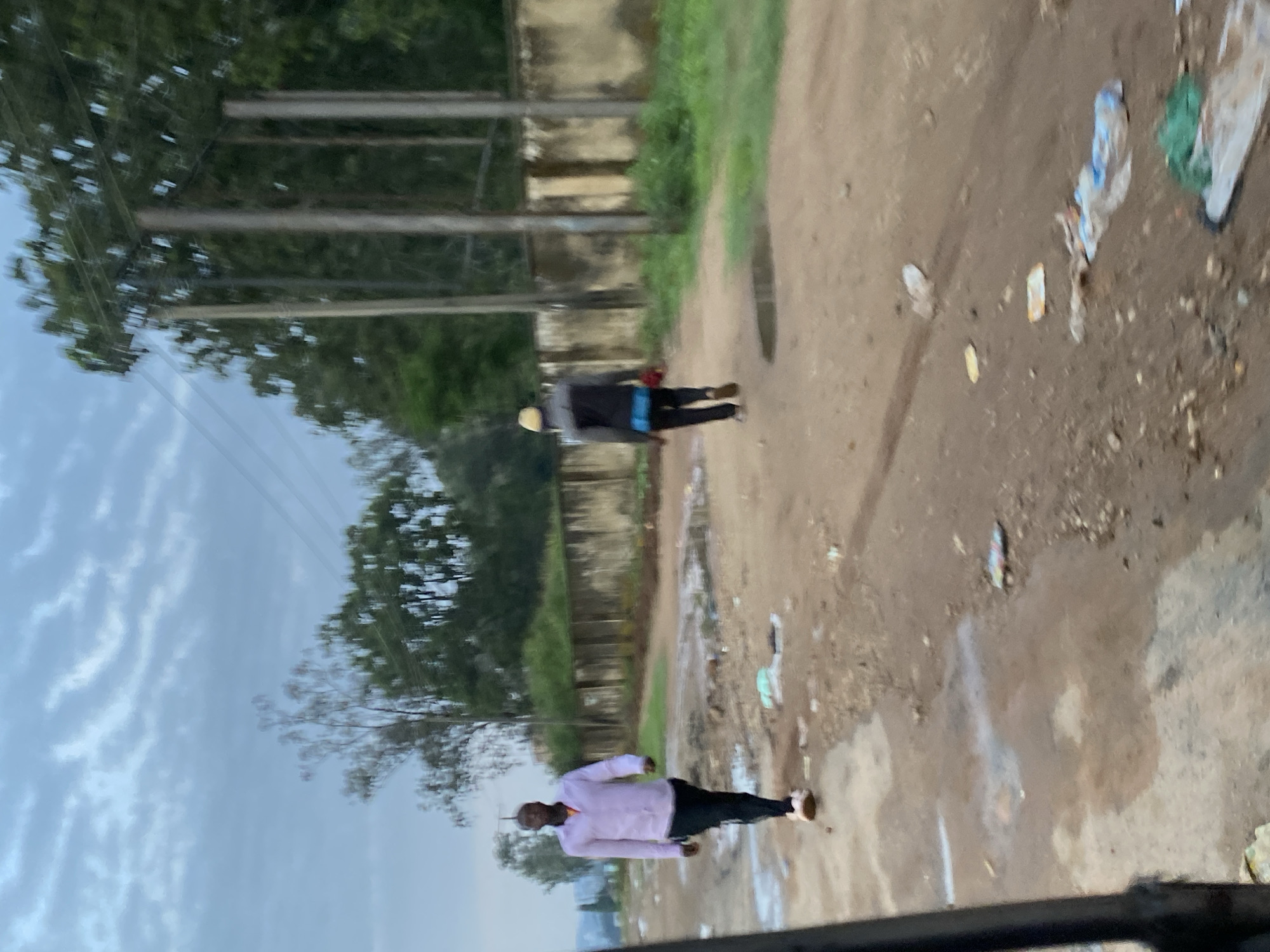 The image depicts an outdoor scene on a dirt road in Gwong, Nigeria. The sky is overcast with textured clouds. On the left side of the road, a man wearing a light purple long-sleeved shirt and black pants walks towards the viewer. He is wearing light-colored sneakers. Further down the road, a second person, viewed from the back, walks away from the viewer. This individual wears a dark jacket, black pants, a yellow hat, and appears to be carrying a red object in their right hand. The road is uneven and appears to have puddles of water, indicative of recent rain. Litter, including plastic bags and other debris, is scattered along the road. Behind the road, a low, weathered concrete wall runs horizontally, with green vegetation and trees beyond it. Utility poles with power lines are also visible in the background. The overall impression is of a somewhat unkempt urban or semi-urban environment under a cloudy sky.