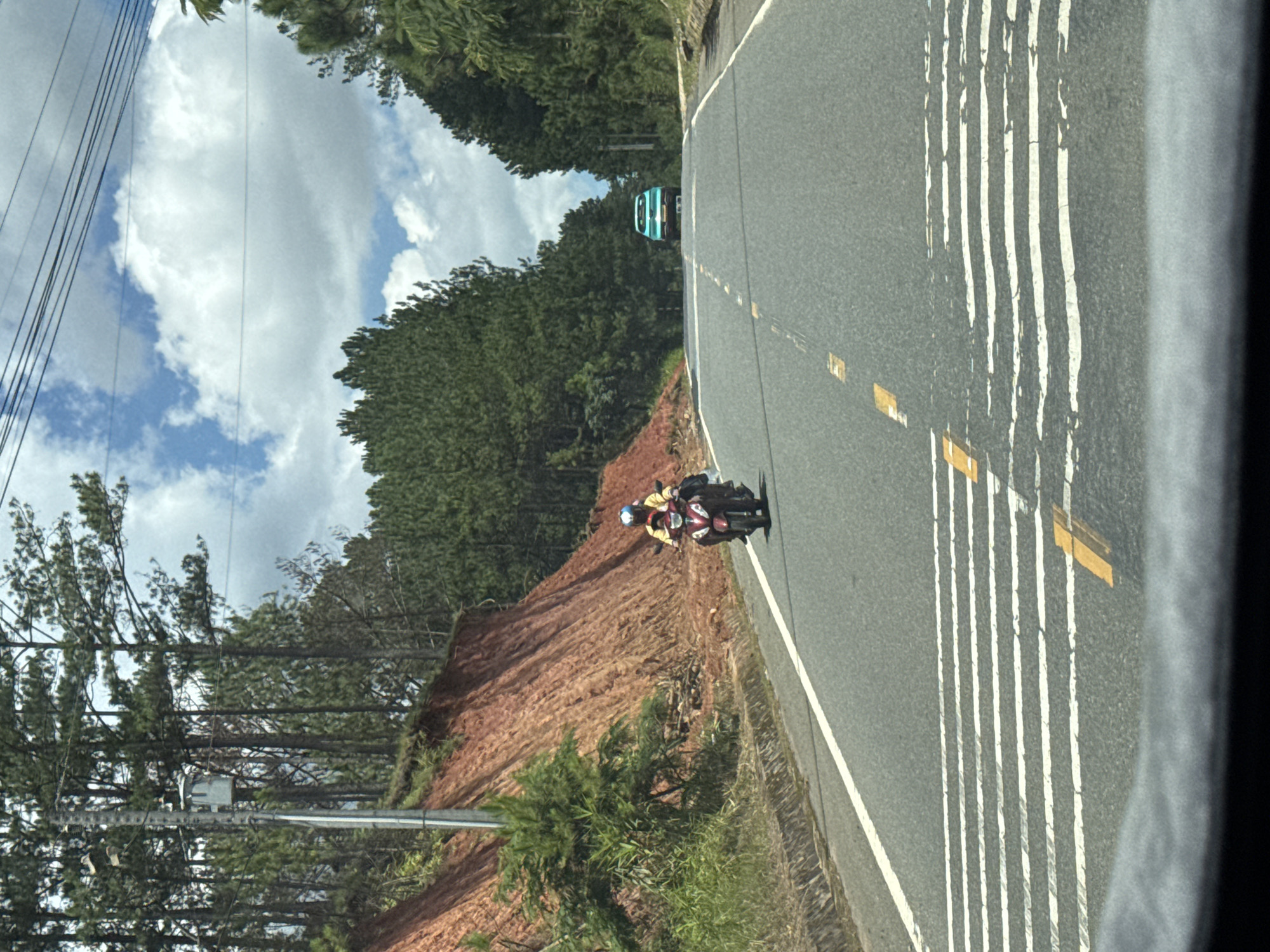 The image depicts a road scene in Thôn 6, Vietnam, characterized by a mountainous terrain with dense pine forests lining the sides of the road. The road itself is paved and appears to be a two-lane highway with white and yellow lane markings. In the foreground, a motorcyclist wearing a yellow shirt and a blue helmet is riding towards the viewer on the right side of the road. The motorcycle is dark red. In the distance, a teal-colored car is also visible, heading away from the viewer. The left side of the road features a steep, eroded embankment with reddish-brown soil, covered in sparse green vegetation and trees. The sky is partly cloudy with bright blue patches and white cumulus clouds. Utility poles and overhead wires are present along the roadside.