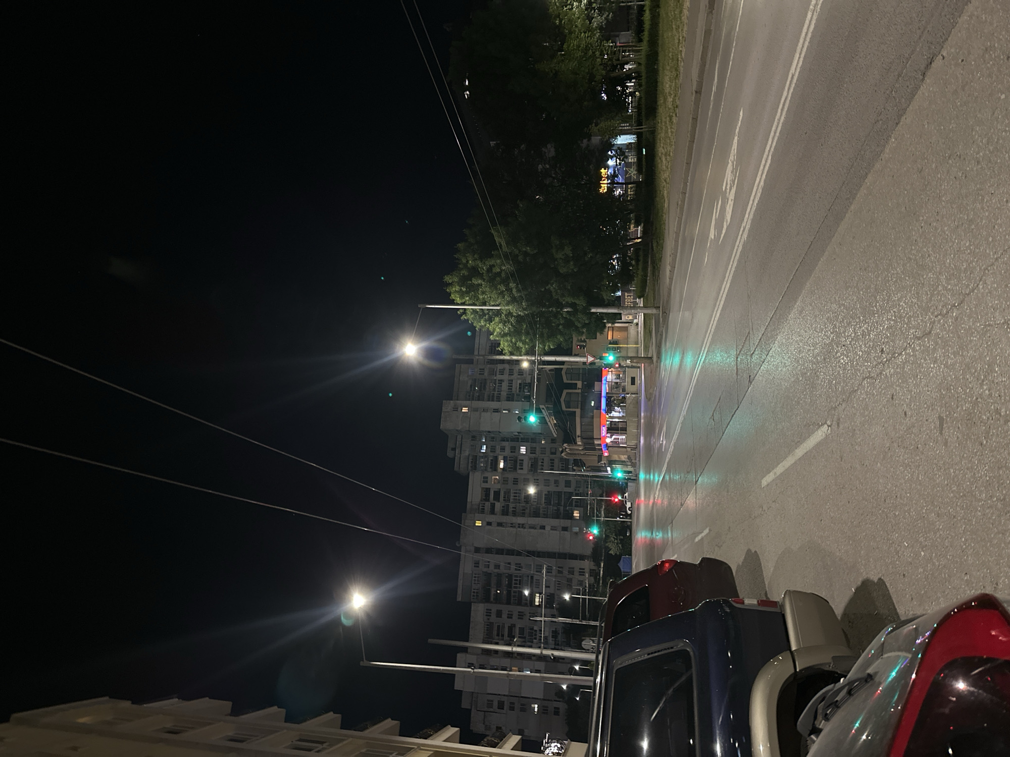 This is a vertical, medium-shot, eye-level, nighttime outdoor photograph taken from a street in an urban environment. The scene is dominated by a street with traffic lights and cars parked on the side. In the foreground, to the left, is the rear of a dark blue SUV, followed by a red SUV and another dark-colored vehicle. The street is wet, reflecting the green lights of a traffic signal visible further down the road. Beyond the street, a row of apartment buildings with lit windows stretches across the background. Trees are visible to the right, with streetlights illuminating the scene. The sky is dark and clear, with visible utility wires overhead. The overall impression is that of a city street at night, possibly after rain.