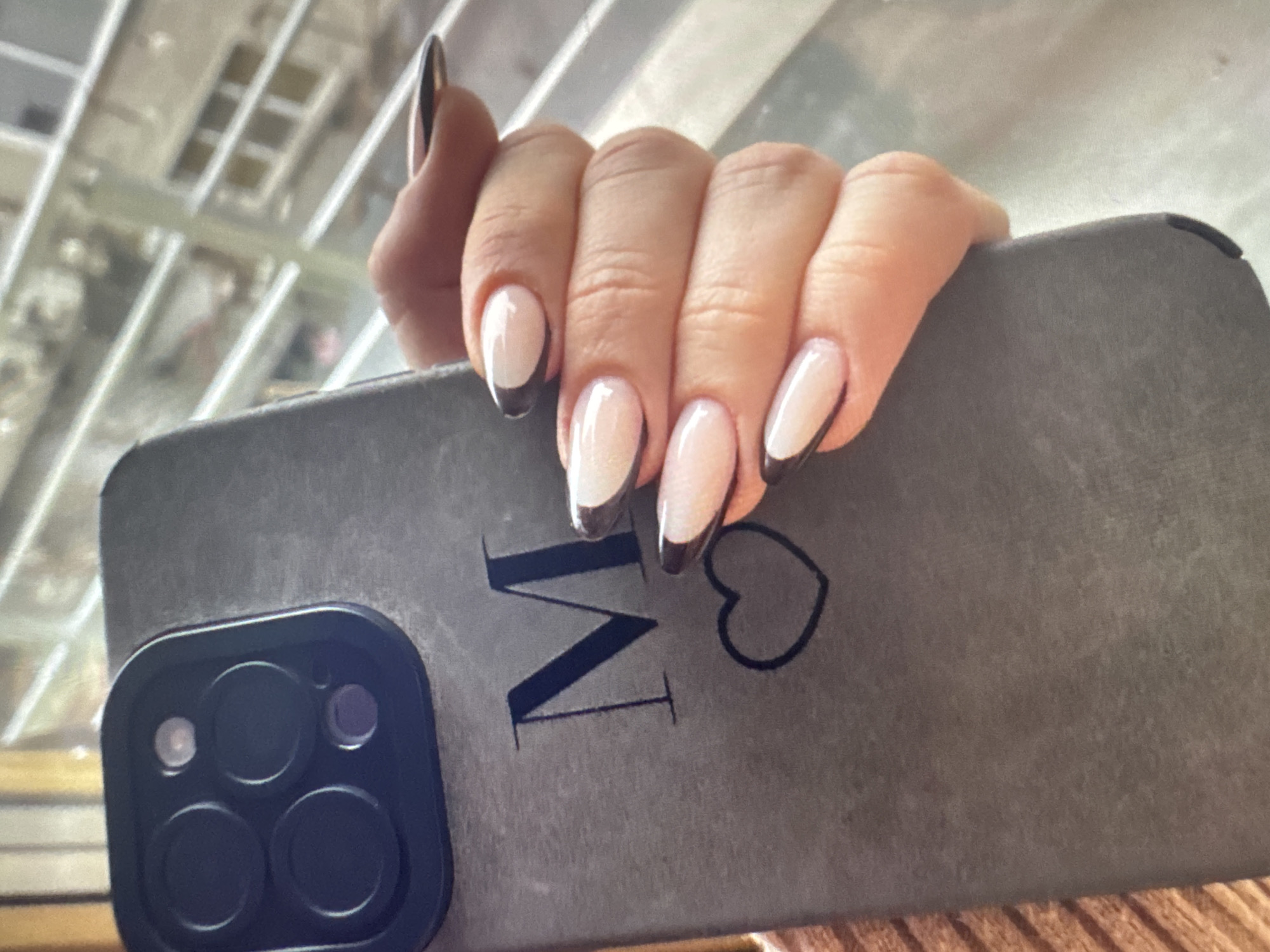 In this close-up, eye-level shot, a person's hand with French manicure nails is holding a dark grey phone case. The phone case has a matte finish and features a dark blue letter "M" and a heart outline. The background shows a blurred view of window blinds and buildings, suggesting an indoor setting, possibly an apartment or office. The overall lighting is natural and diffused. No specific location in Plovdiv, Bulgaria, can be identified from the visual information provided.