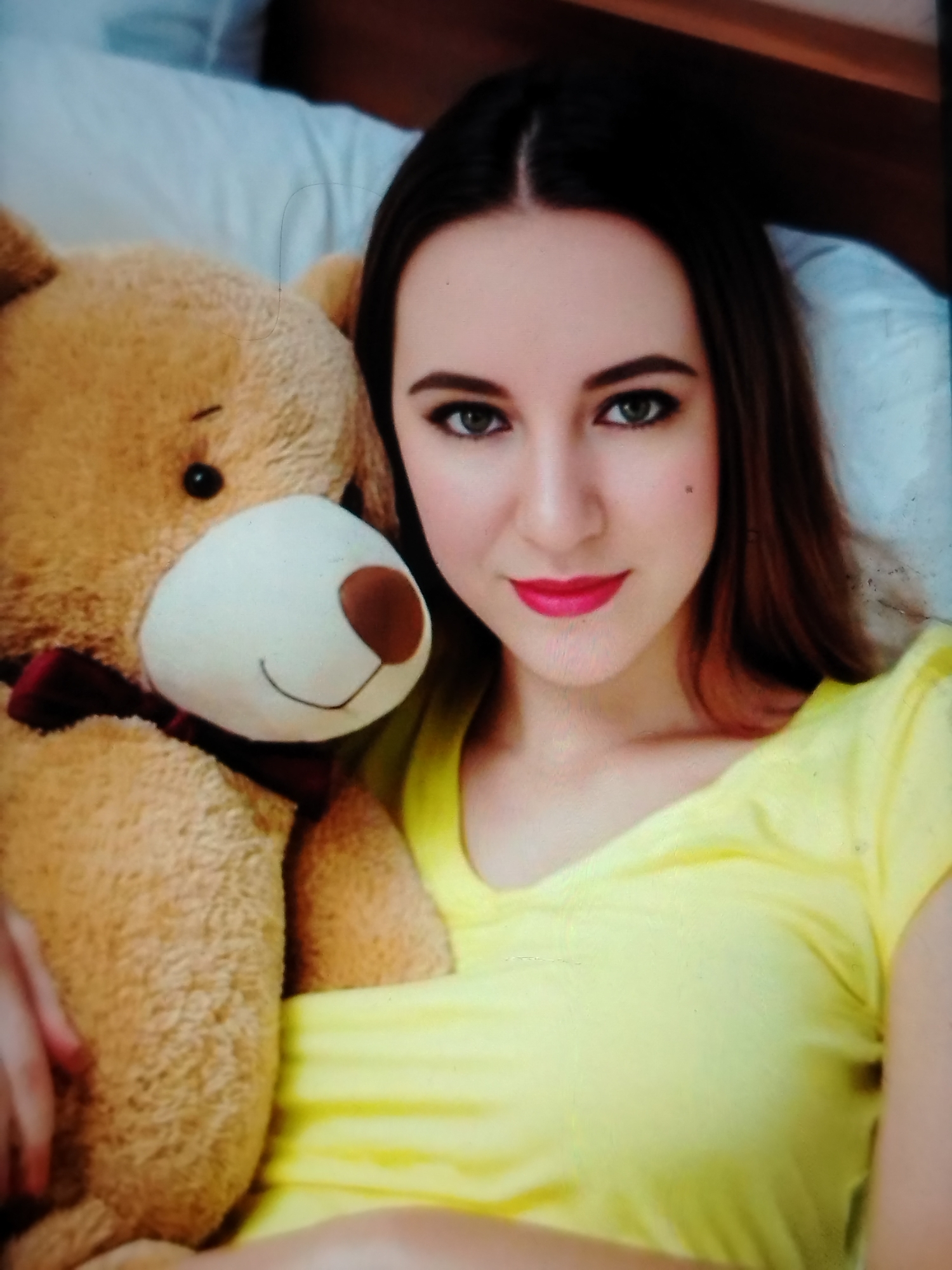 The image captures a young woman posing with a large teddy bear in what appears to be an indoor setting, likely a bedroom. The woman, with long dark hair parted down the middle, is looking directly at the camera. She has green eyes, defined with eyeliner, and is wearing bright red lipstick. A small dark mole is visible on her right cheek. She is wearing a simple yellow t-shirt. Her expression is calm and direct.
The teddy bear, a prominent subject, is large, light brown, and fluffy, positioned to the left of the woman and partly in front of her. It features a white snout with a brown nose and a drawn-on smile. A dark red bow tie is visible around its neck. One of the woman's hands is resting gently on the bear.
The background is softly blurred, showing elements of a bed, including white bedding or pillows, and a wooden headboard or furniture. There is no visible text in the image. The general atmosphere is relaxed and personal. The provided location context for this image is Zhlobin, Belarus.