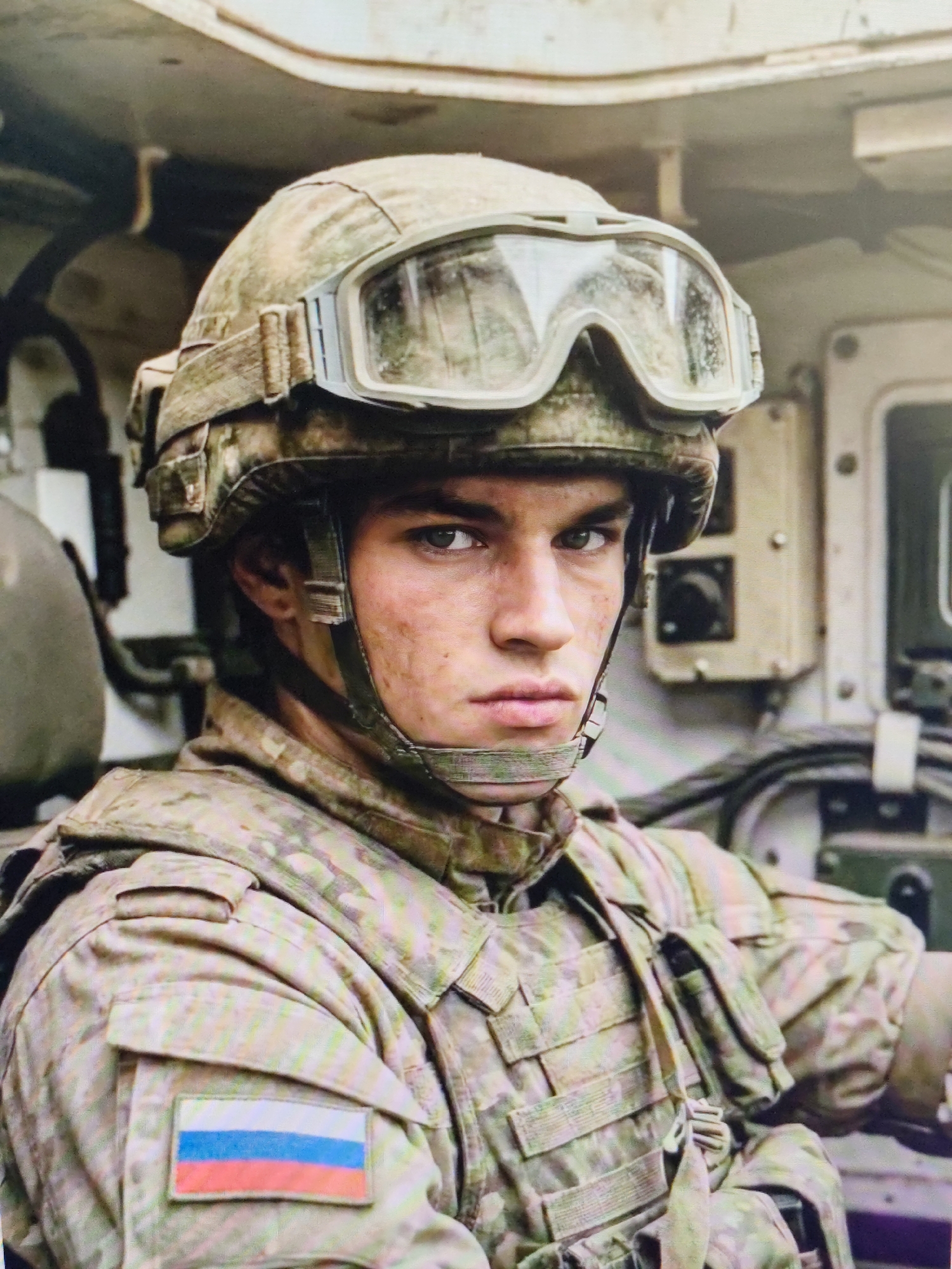 A close-up, head-and-shoulders portrait shows a young male soldier wearing a camouflage helmet with goggles and a camouflage uniform. He is in the foreground, facing the camera. His face is smudged with dirt, and he has a serious expression. On his left shoulder, a Russian flag patch is visible. The background appears to be the interior of a vehicle, with various metallic and electrical components. The scene is not identifiable as being in the city of Syktyvkar, Russia.
