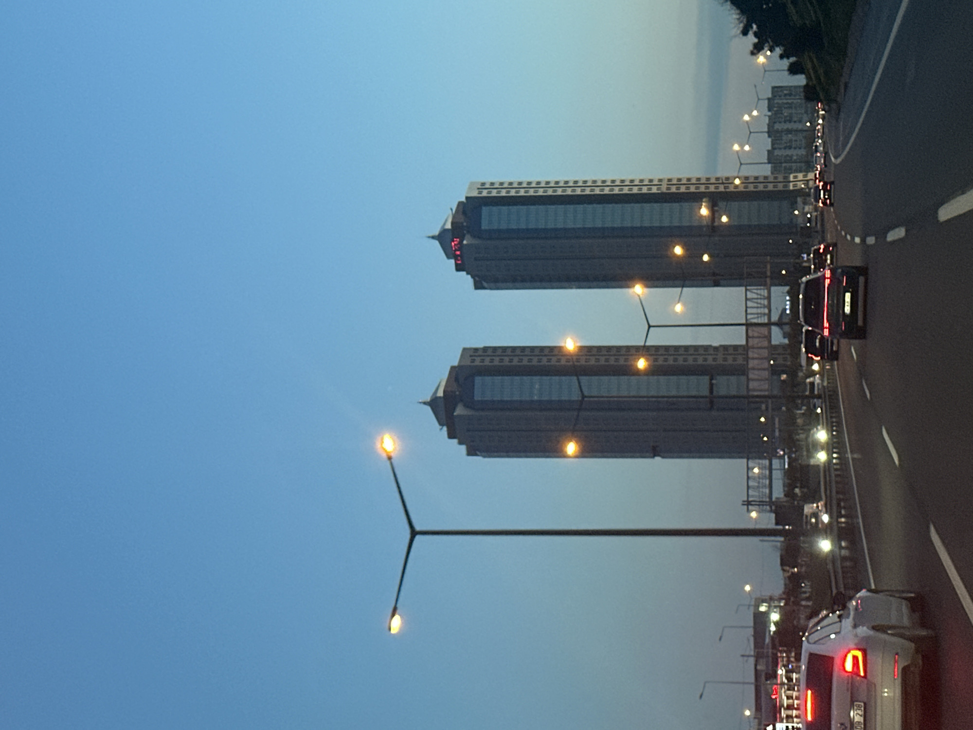 The image depicts a view from a road in Esenler, Turkey, featuring two high-rise buildings. These buildings have a similar design, with a multi-story structure and a pointed spire at the top. Each building also has a clock near the spire. The photo was taken at dusk or dawn, as indicated by the blue sky and the lit streetlights. The road appears to be a highway, with multiple lanes and cars driving on it. There are streetlights along the side of the road. The buildings and road are typical of an urban environment in Esenler.
