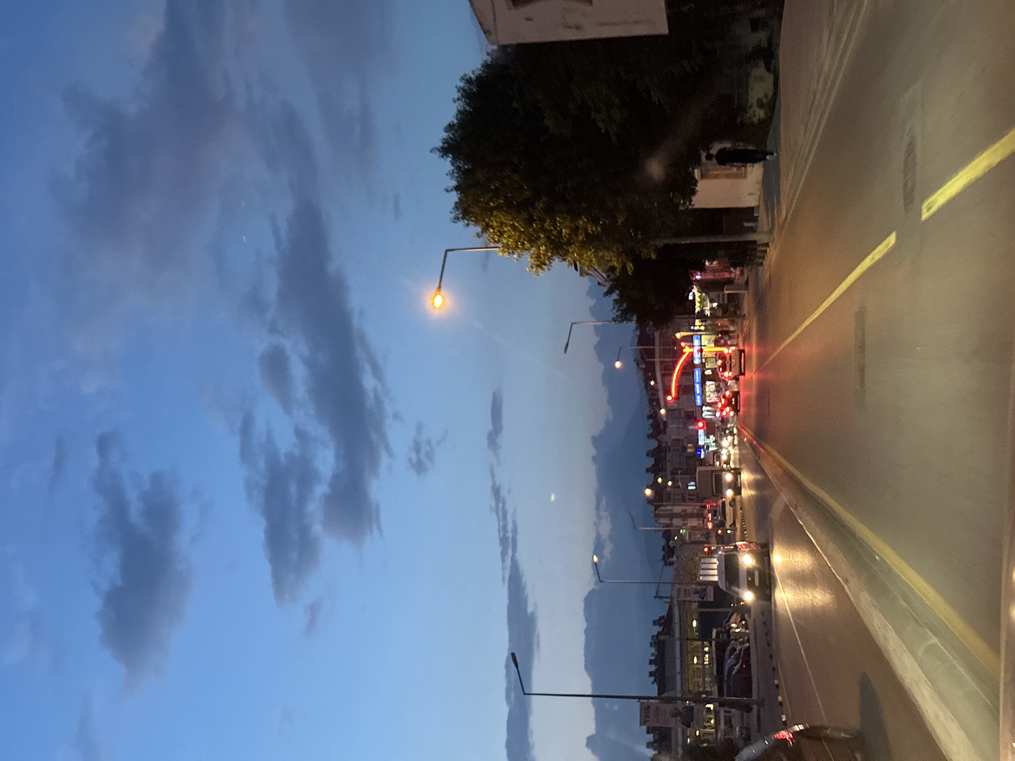 The image depicts a street scene in Fethiye, Turkey, at dusk. The view is oriented down a road, with a yellow line marking the center. Streetlights illuminate the roadway, and buildings line the sides of the road. There is traffic, including cars and other vehicles. A tree is on the side of the road on the left, and the sky above is blue with clouds.