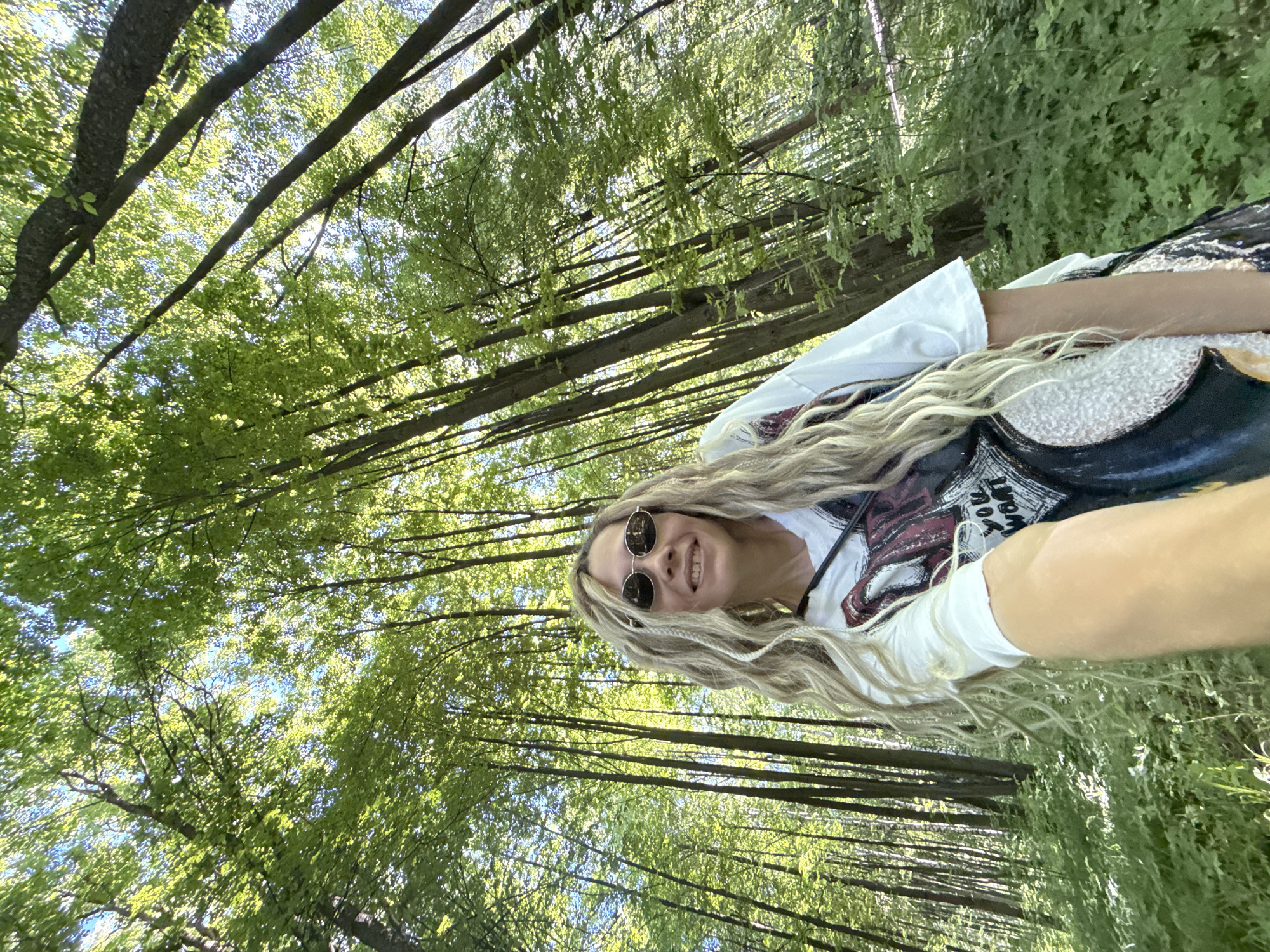 The image depicts a female individual taking a selfie in a dense forest located in Rusanovo, Russia. The woman is positioned in the lower-right quadrant of the frame, holding the camera with an extended arm. She has long, wavy, light-colored hair, some sections of which are braided. She wears round sunglasses with dark lenses and thin metallic frames. Her expression is a smile, revealing a gap between her two front upper teeth. She is wearing a white t-shirt featuring a dark graphic design, part of which reads "YOU WANT." A dark strap is visible around her neck. The background consists of numerous tall, slender trees with vibrant green leaves, suggesting a lush deciduous forest. Sunlight filters through the canopy, creating bright highlights on the foliage and a visible blue sky through gaps in the leaves. The forest floor is covered with green undergrowth.